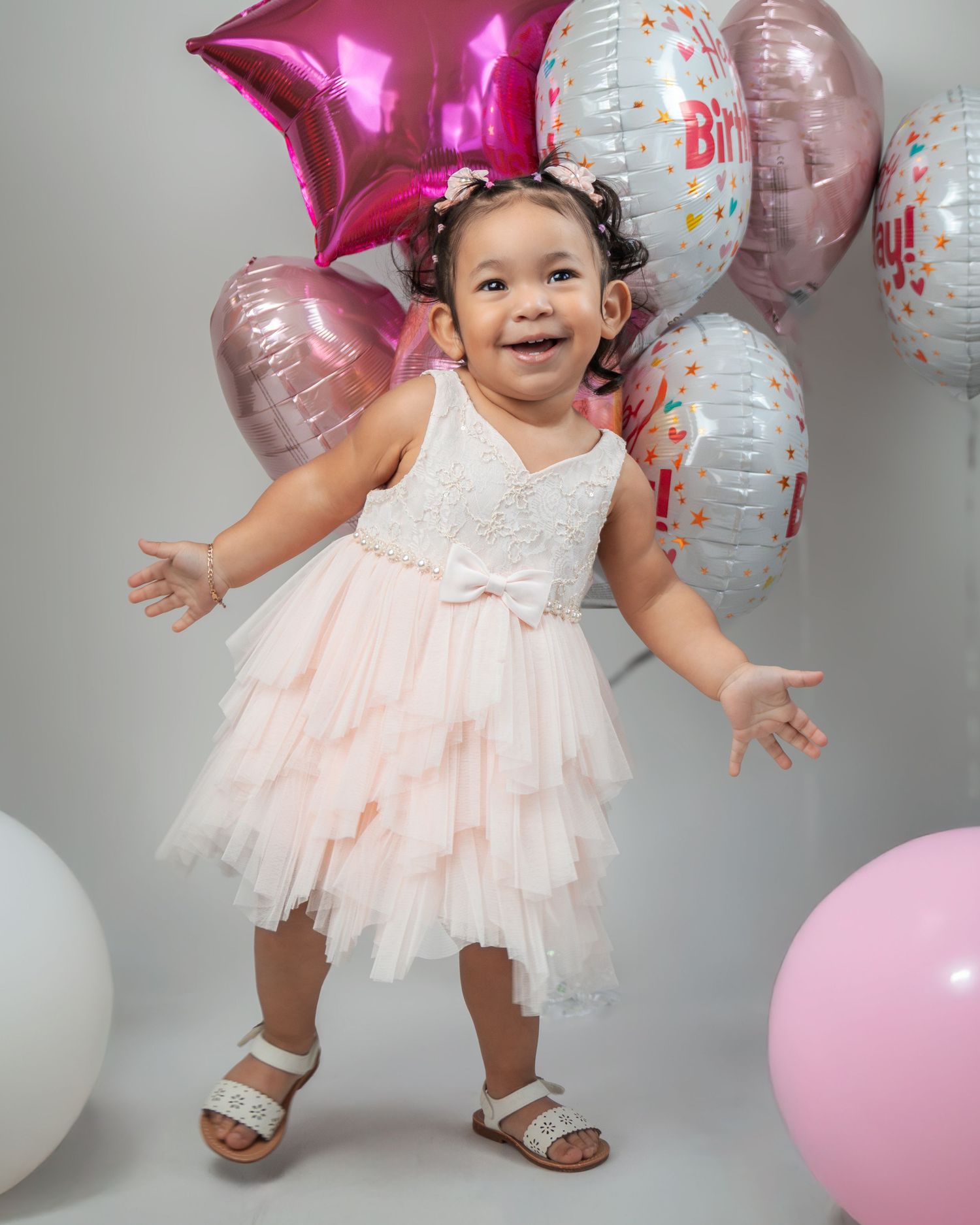 A small child in a pink tulle dress poses joyfully against a backdrop of pink and silver birthday balloons.