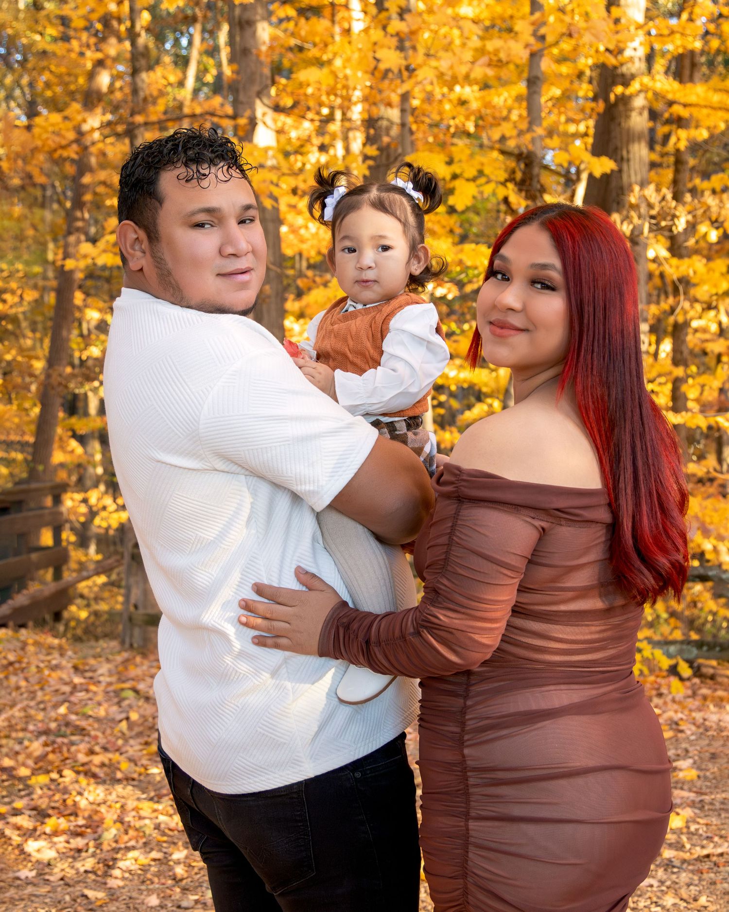 A young family poses outdoors in autumn, man in white shirt holding toddler, woman in brown off-shoulder dress, fall foliage background.