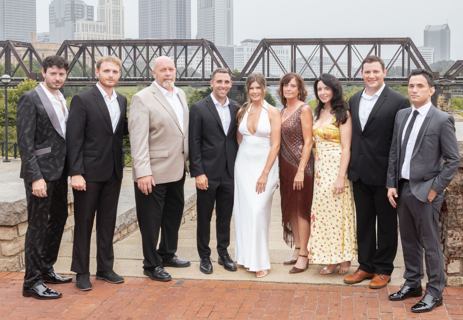 Group of nine elegantly dressed people posing outdoors near a river bridge in an urban setting.