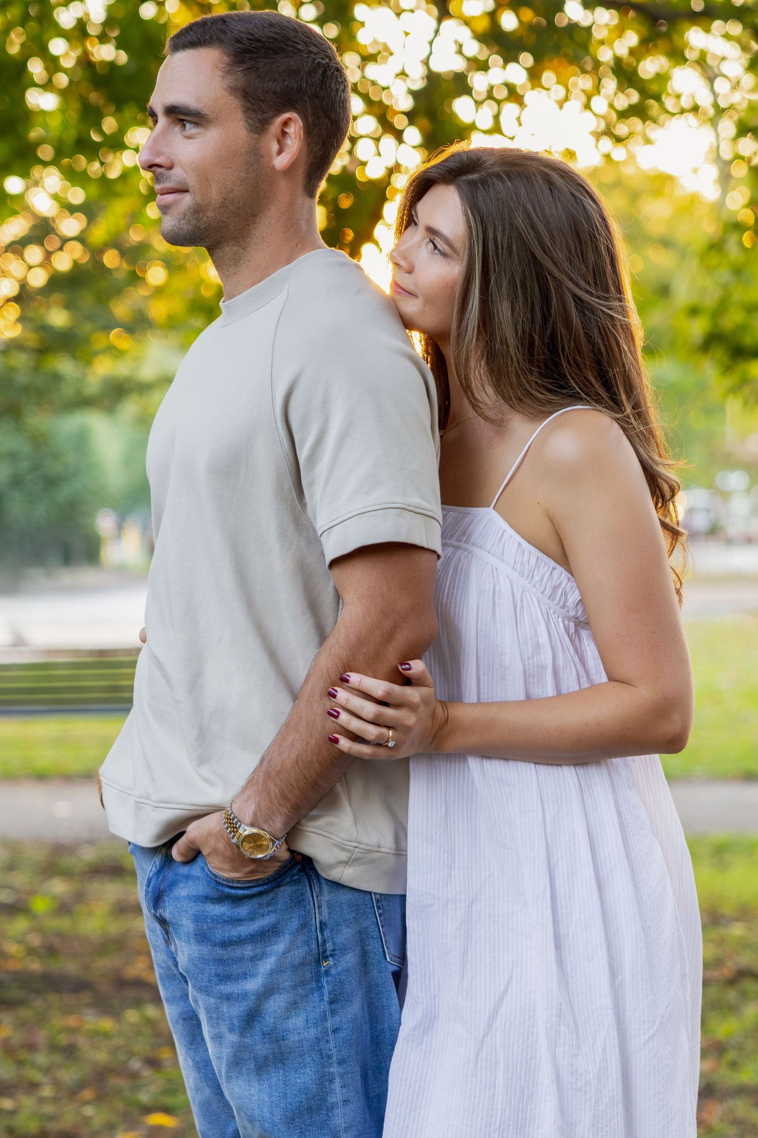 A couple stands together outdoors, woman in white dress embracing man in gray shirt and jeans in a sunny park setting.