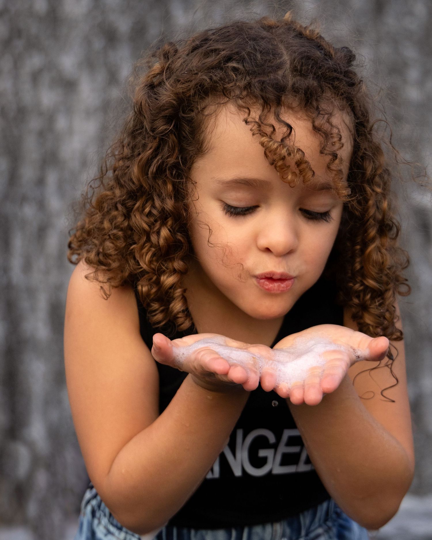 A young girl with curly hair blows glitter from her cupped hands, wearing a black tank top outdoors.