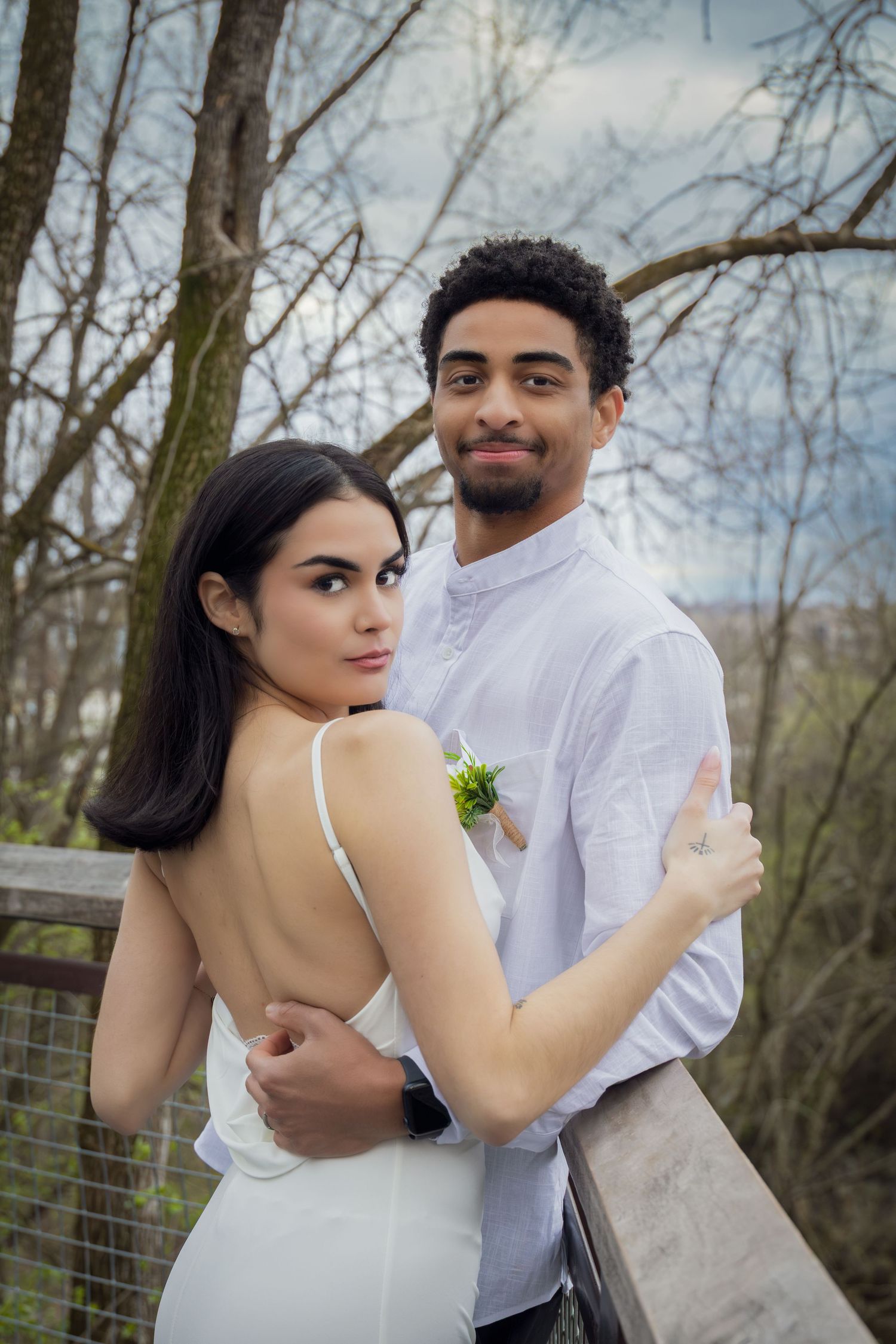 A couple in white attire embraces on a wooden bridge surrounded by bare trees, posing for a romantic outdoor portrait.