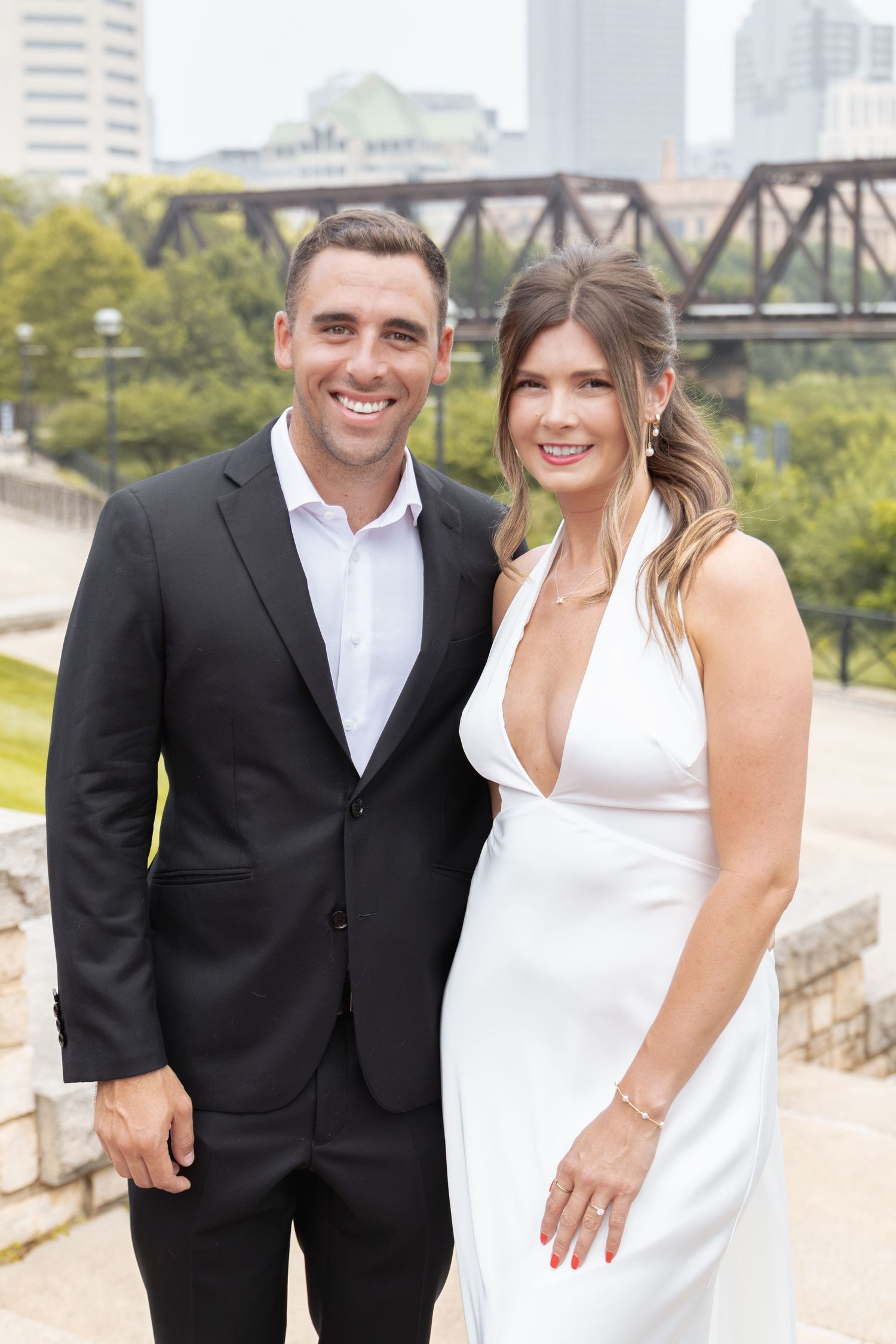 A smiling couple poses outdoors, man in black suit, woman in white halter dress, with a bridge and city skyline behind them.