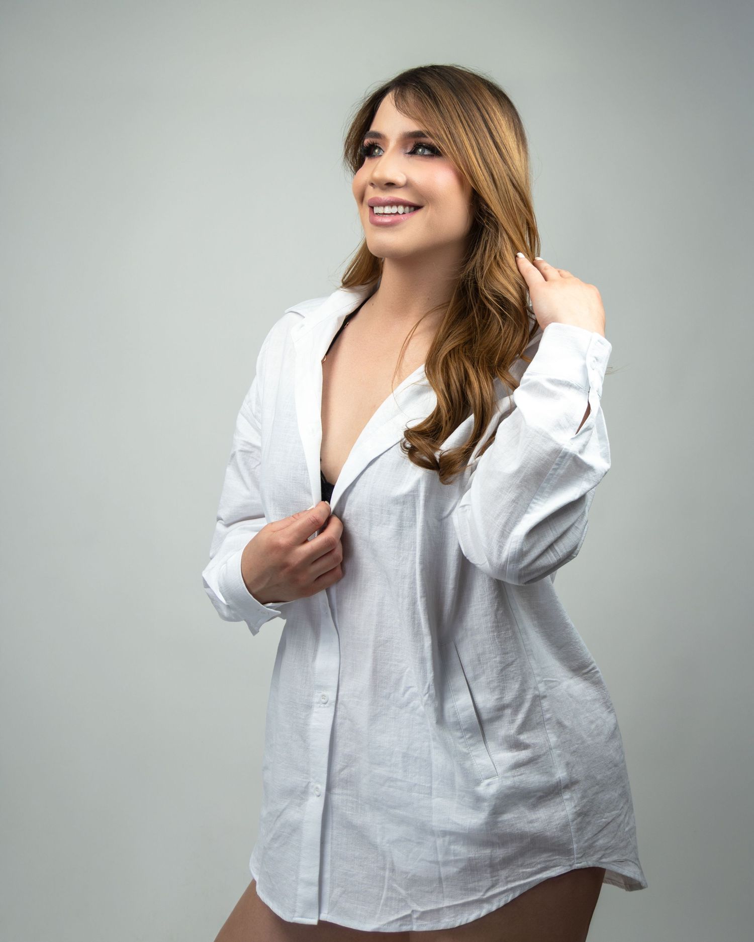 Smiling woman in an open white oversized button-up shirt posing confidently against a light gray background.