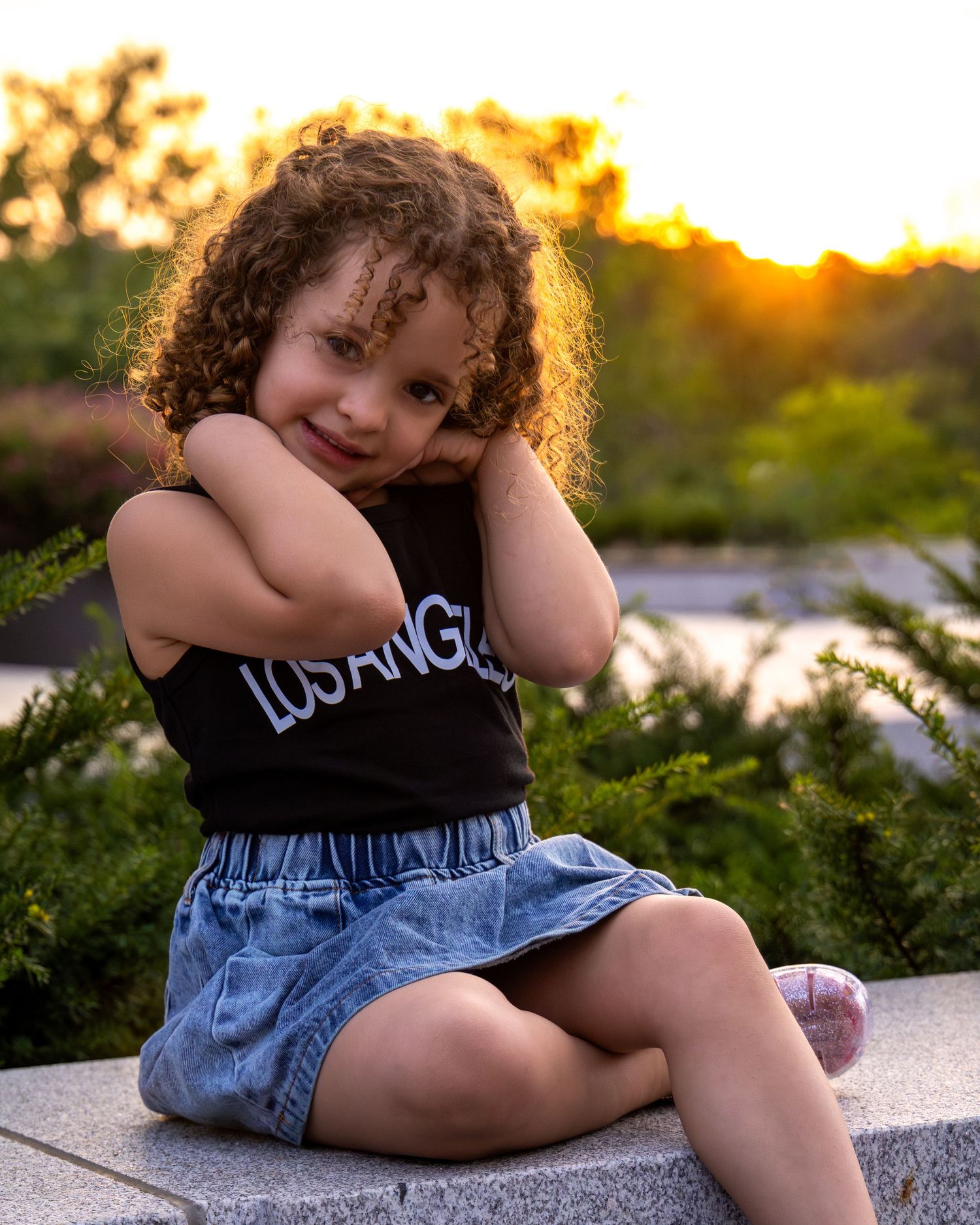Curly-haired toddler girl in black Los Angeles top and denim skirt sitting outdoors at golden sunset.