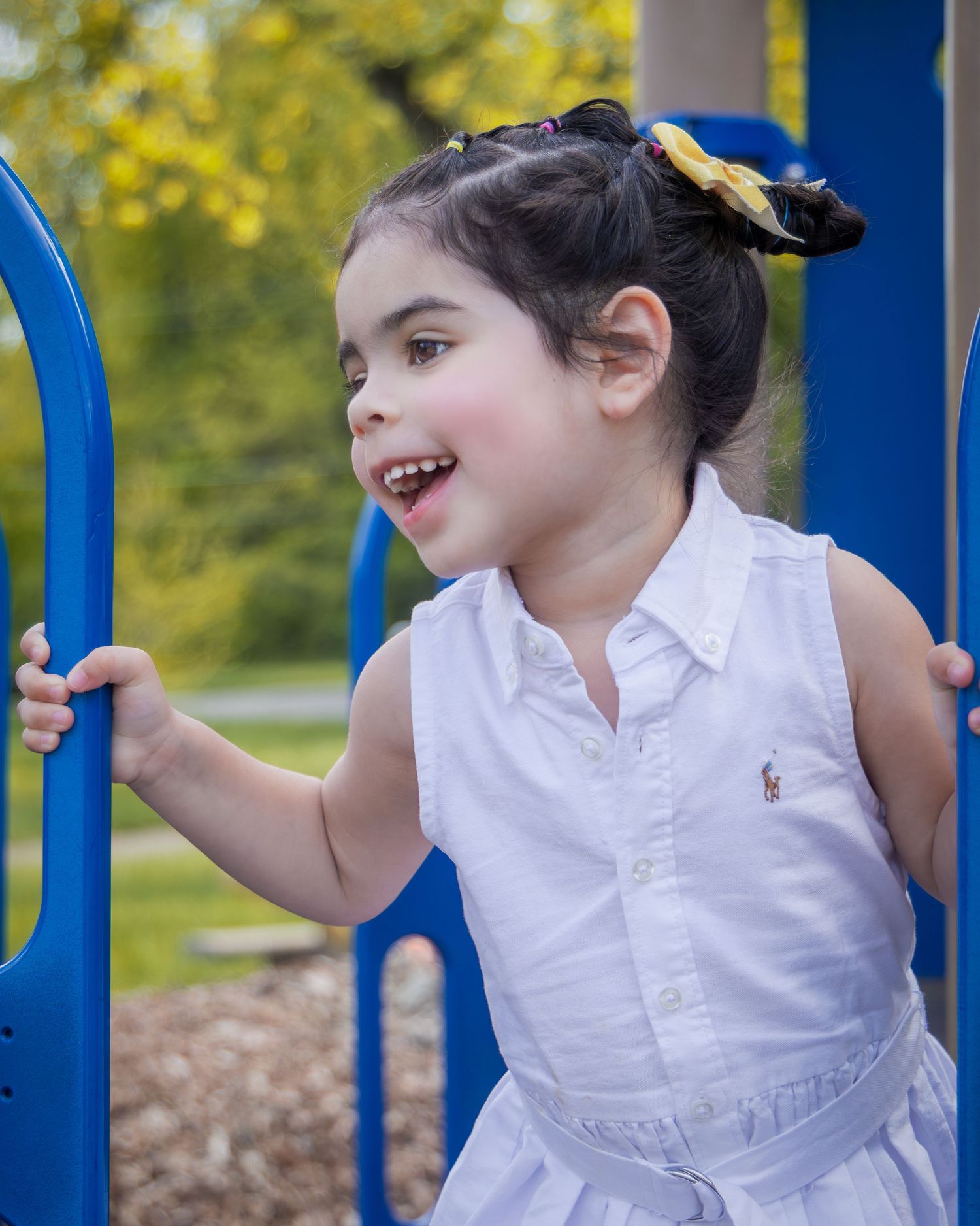 A joyful young girl in a white Ralph Lauren dress plays on blue playground equipment outdoors.