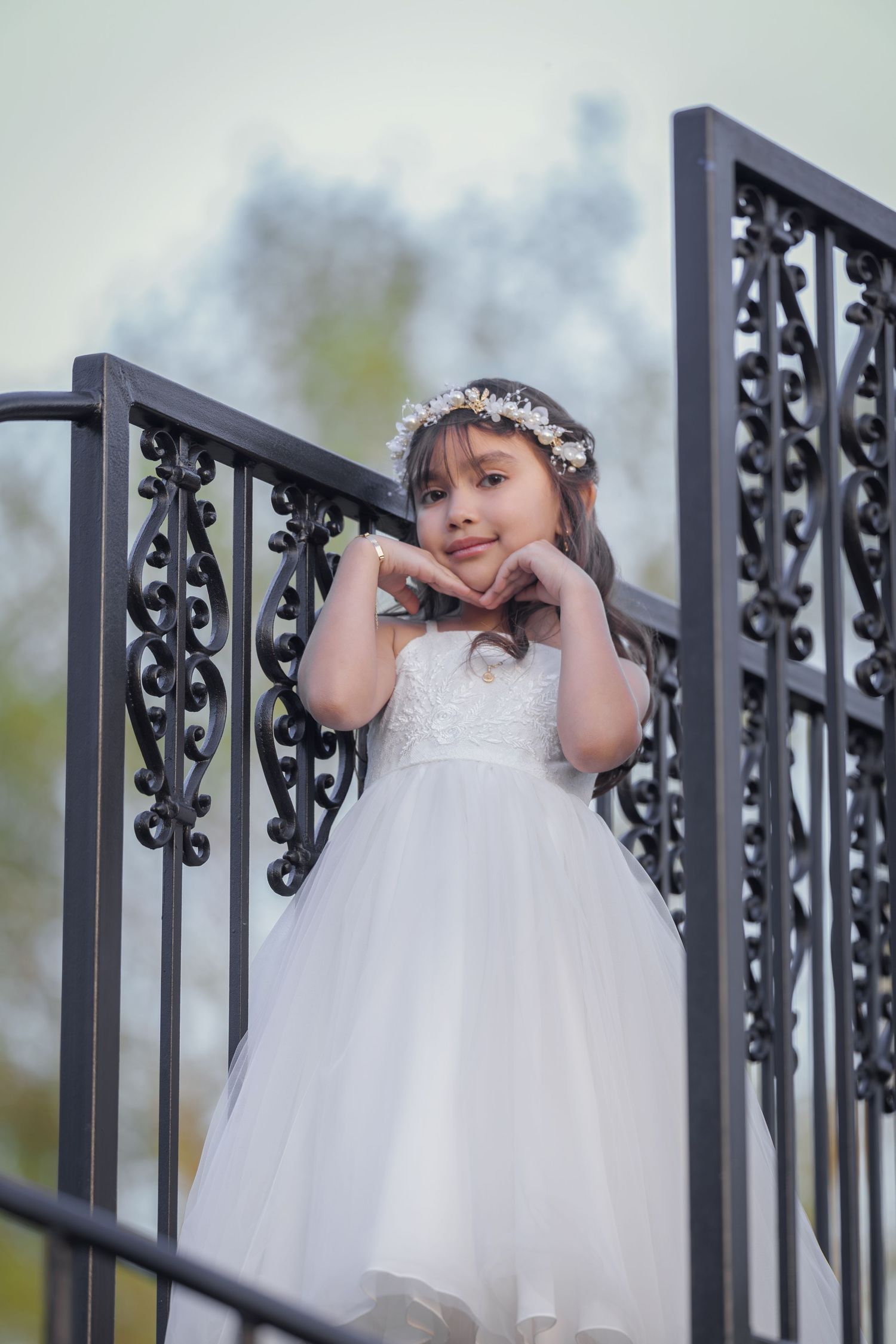 A young girl in a white tulle dress and floral crown poses gracefully on ornate black iron stairs outdoors.