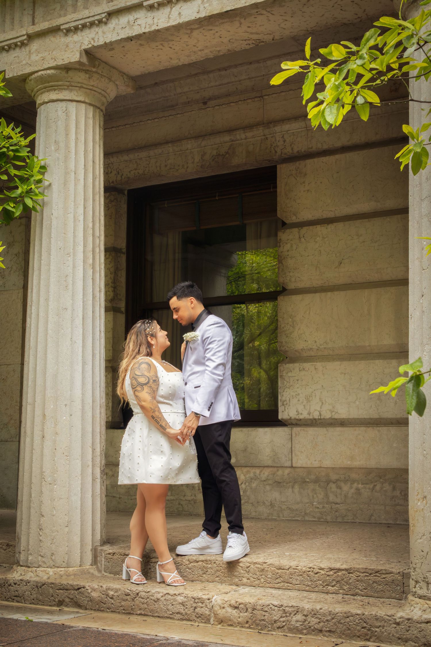 Couple in wedding attire embracing on stone steps between classical columns, bride in short white dress, groom in light suit.