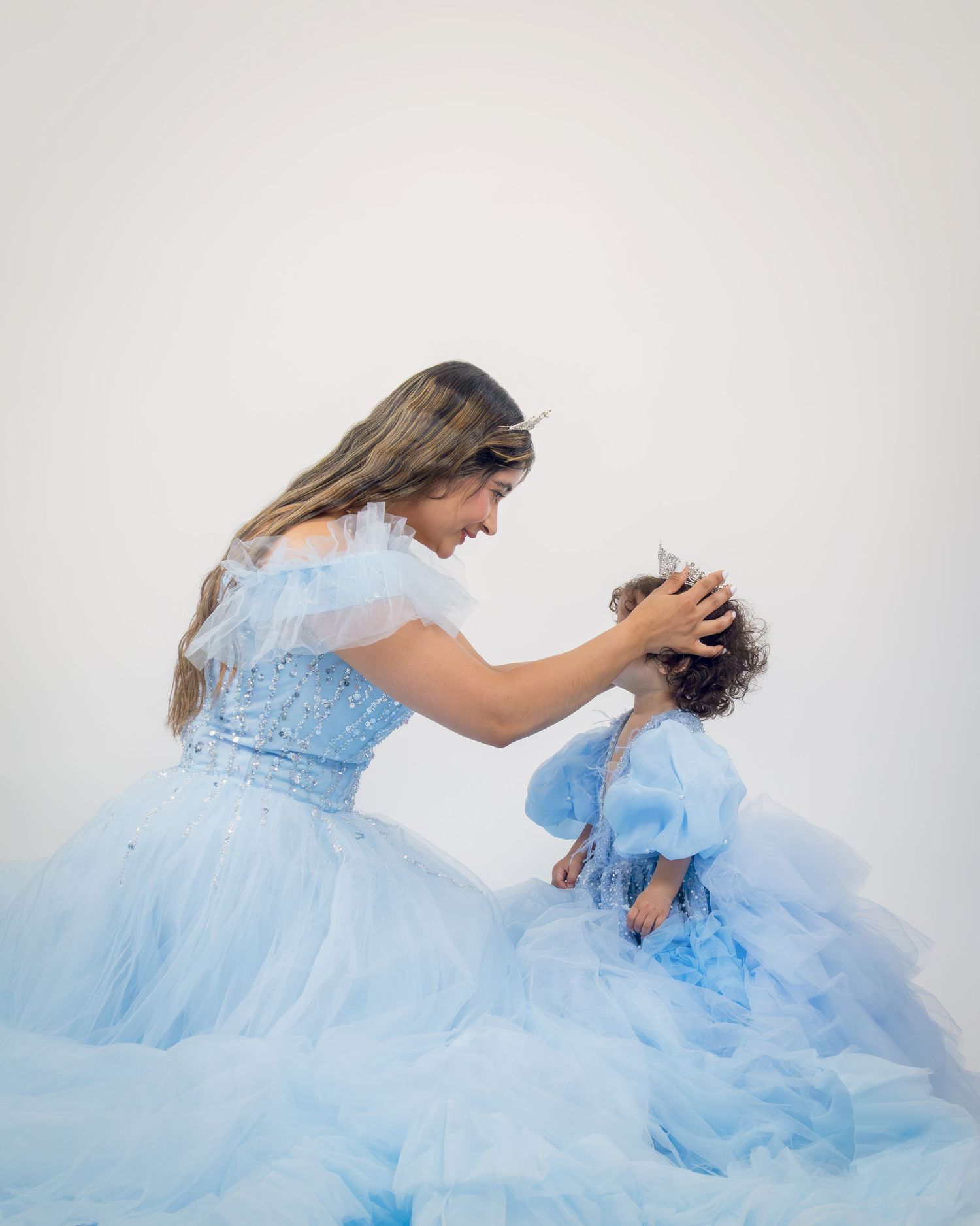 Woman and toddler in matching light blue ball gowns share a tender moment in an elegant photo session.