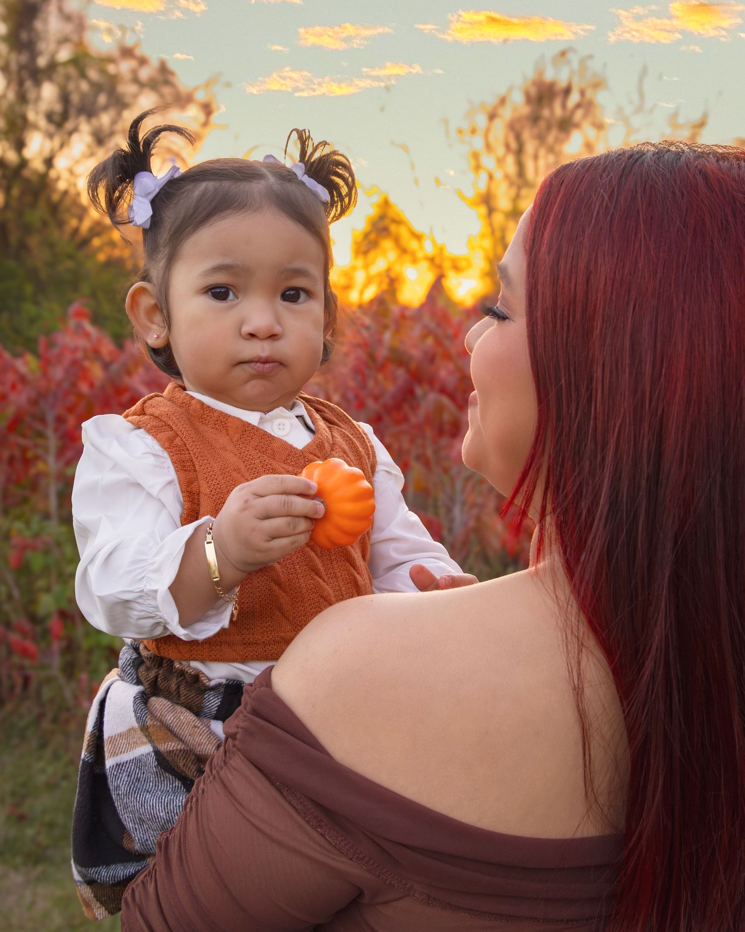 A toddler in an orange dress holds a small pumpkin while being carried by a red-haired woman during autumn sunset.