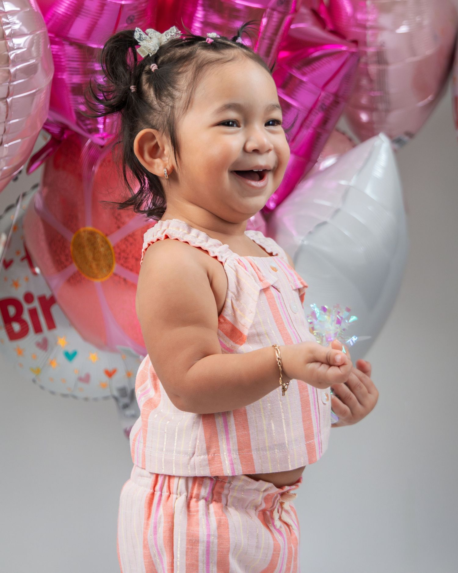 A laughing child in a pink and white striped outfit stands surrounded by festive birthday balloons and decorations.