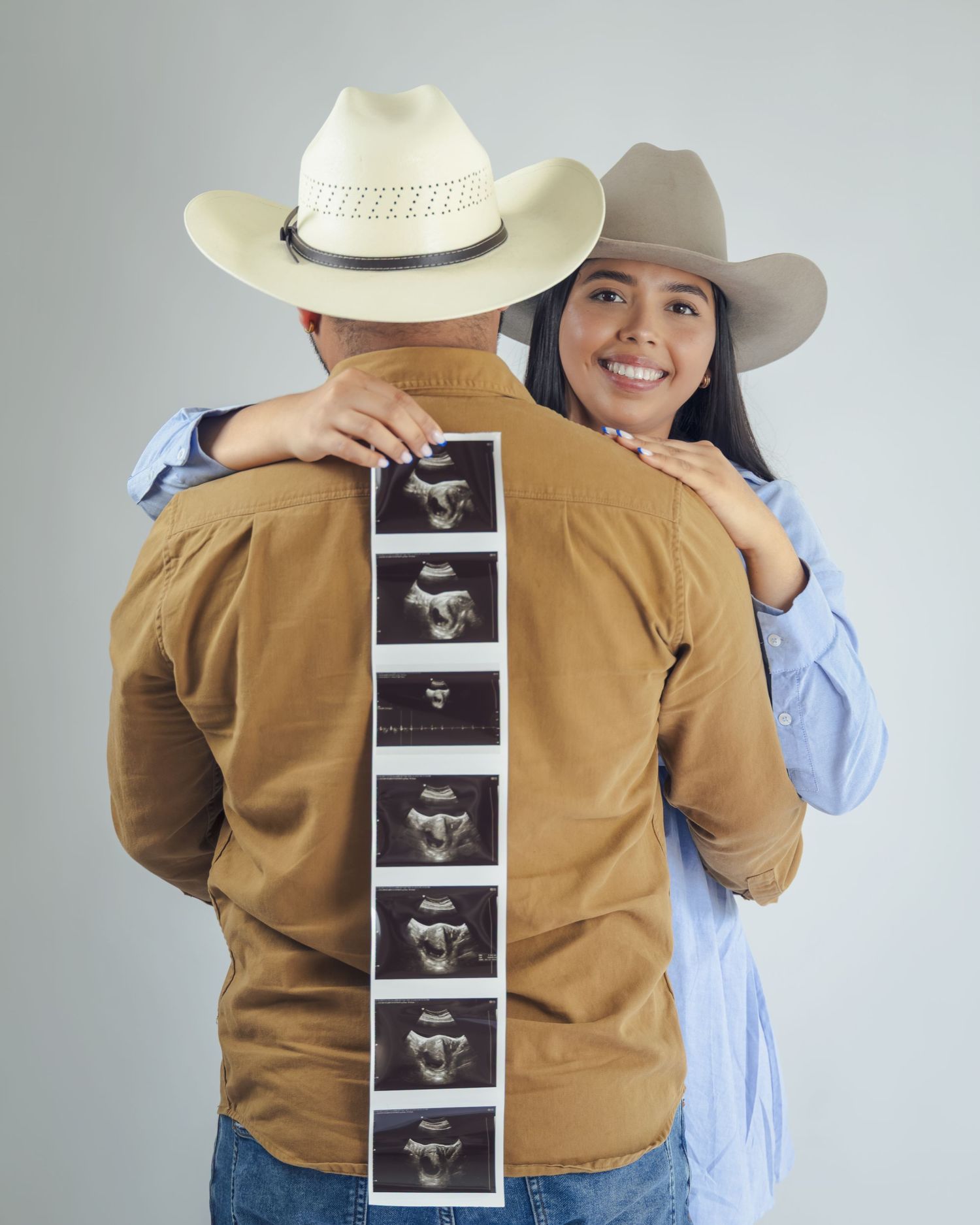 Couple in cowboy hats holding ultrasound photos on man's back, announcing pregnancy in a western-themed photoshoot.