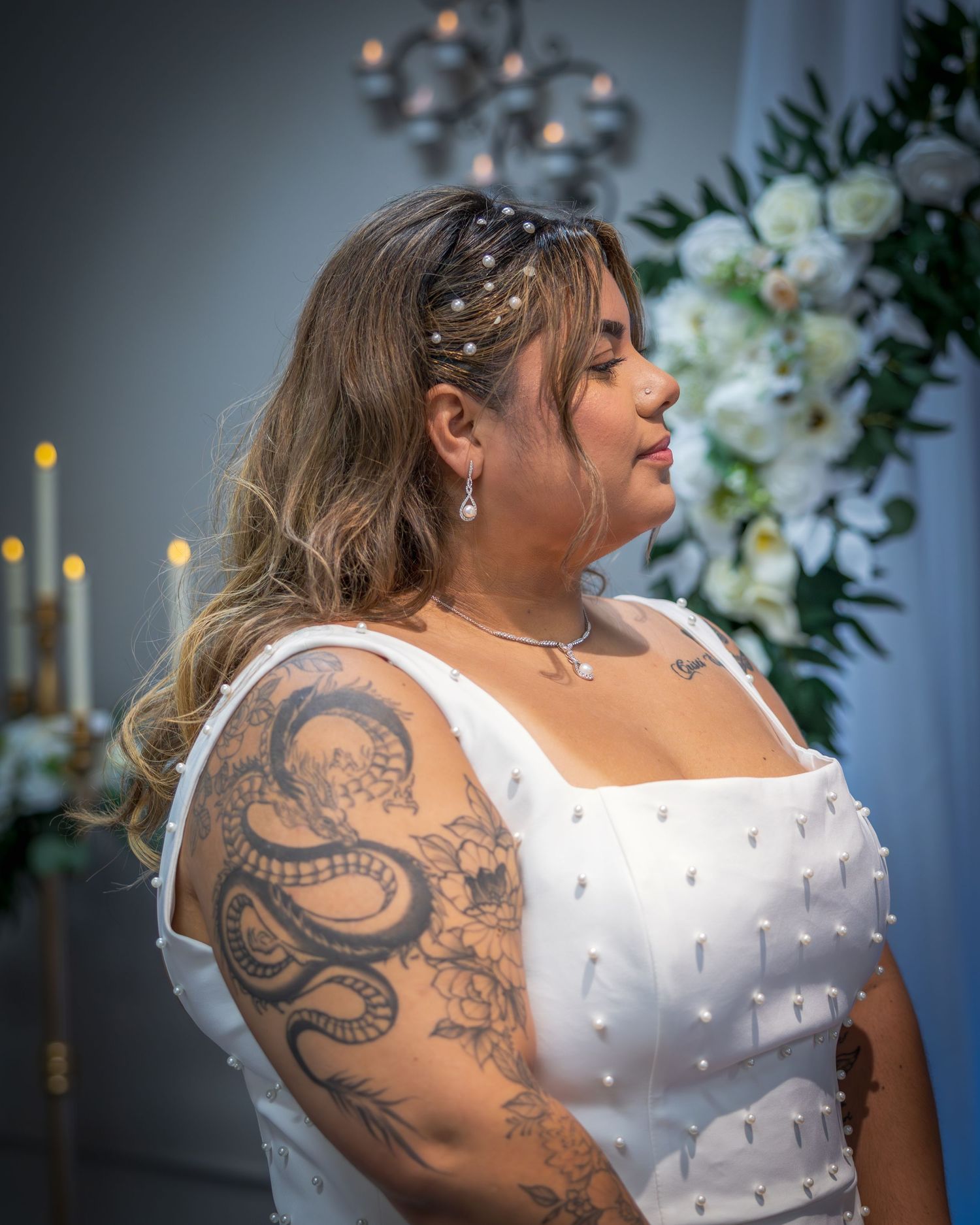 Bride in white dress with snake arm tattoo and pearl hair accessories stands before floral arrangement and candles.