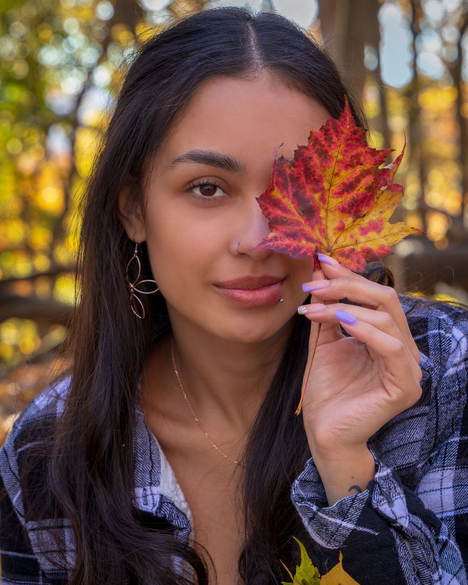 Young woman holding a vibrant red autumn maple leaf over one eye in a sunlit fall forest setting.