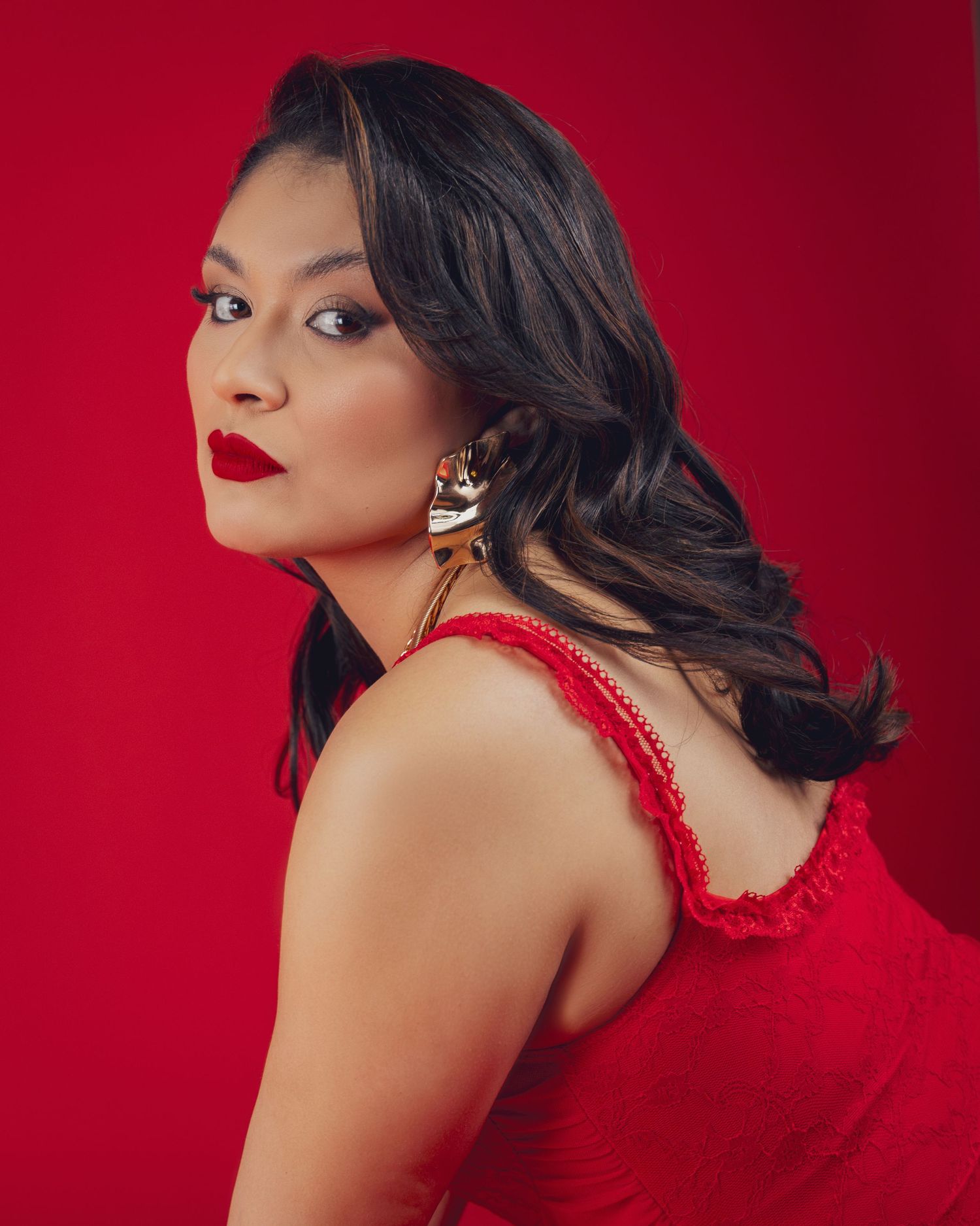Woman in red dress with bold red lips and statement earrings posing against a vibrant red background.