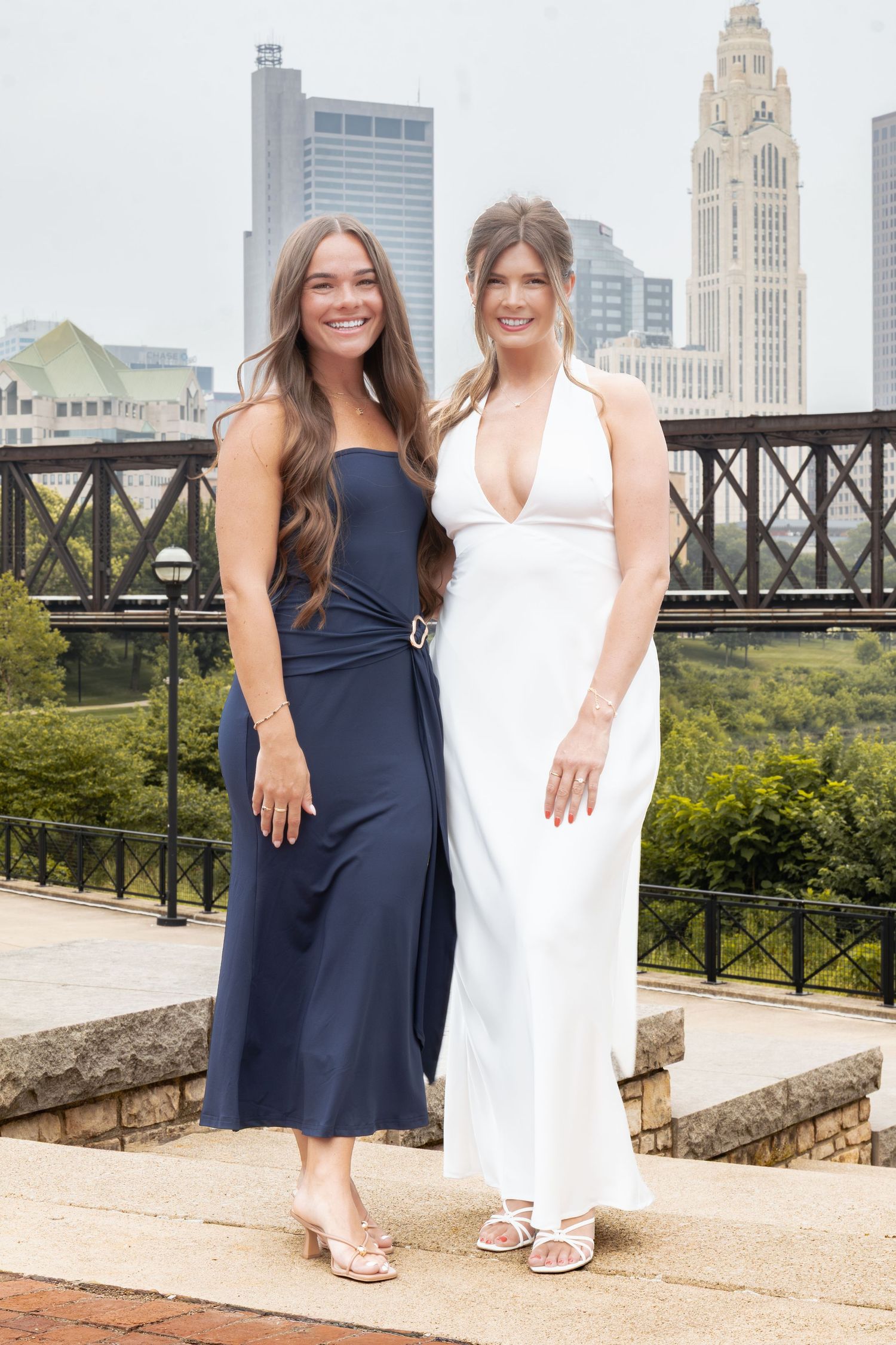 Two women in elegant navy and white gowns pose outdoors with a city skyline backdrop.