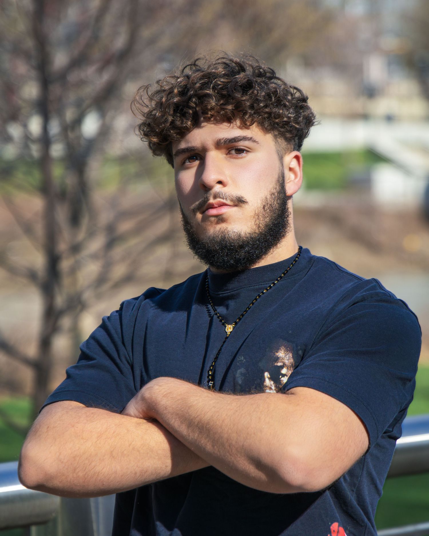 Young man with curly hair and beard, arms crossed, wearing navy blue t-shirt, standing outdoors.