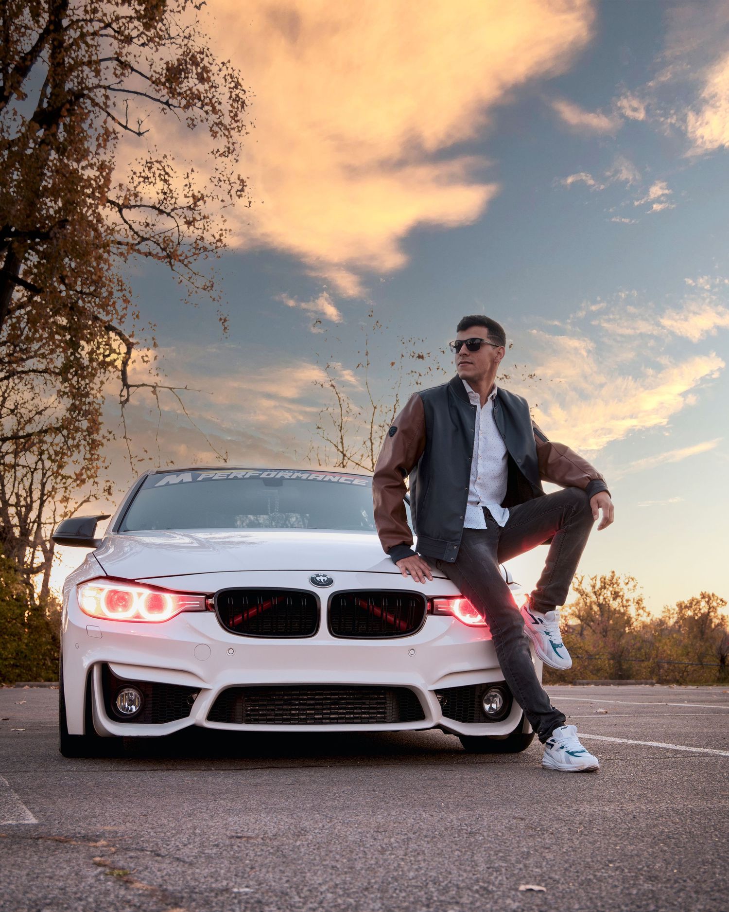Man in sunglasses and brown jacket poses confidently against a white BMW at sunset in a parking lot.
