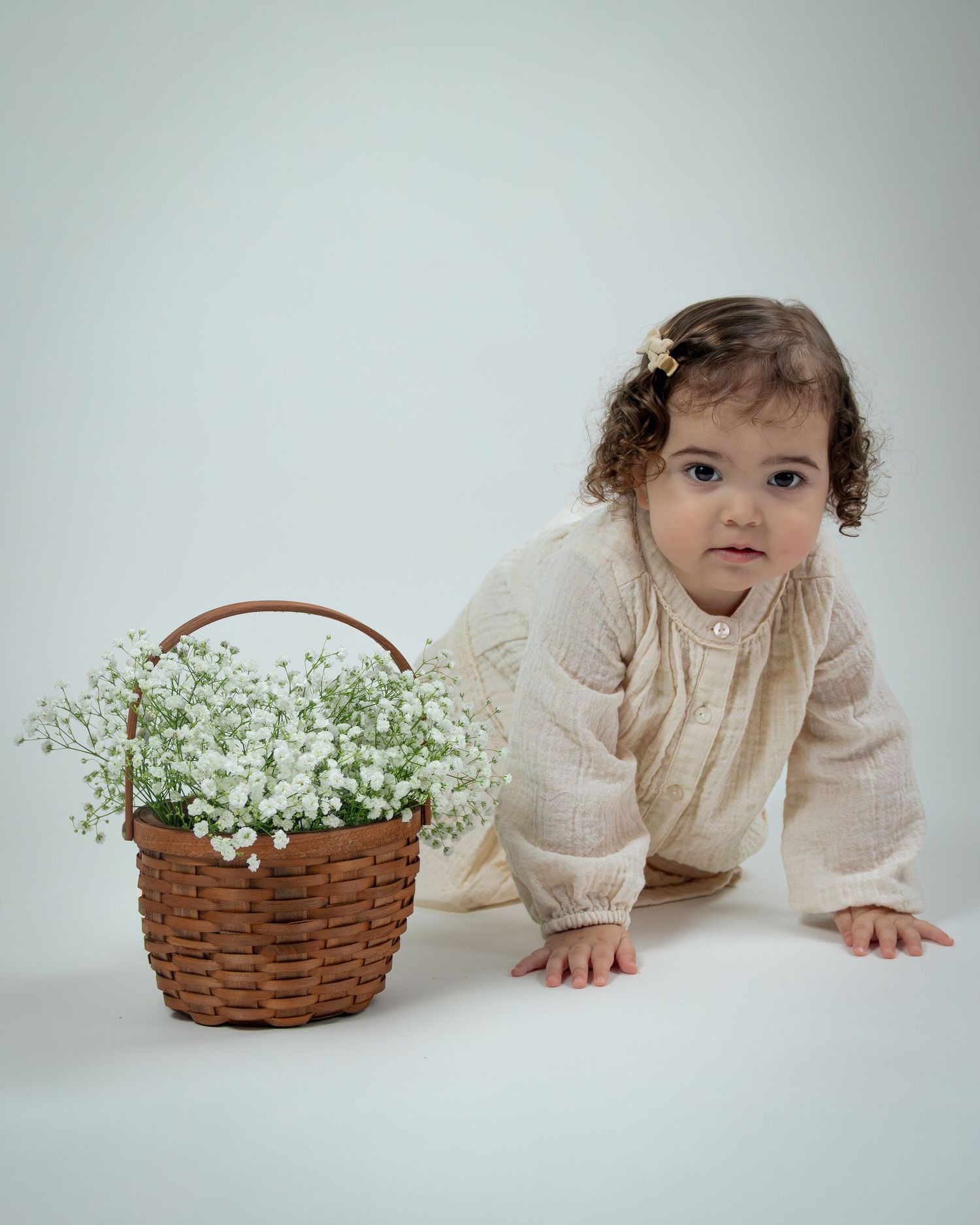 A small child in a cream-colored sweater crawls next to a wicker basket filled with baby's breath flowers on white background.
