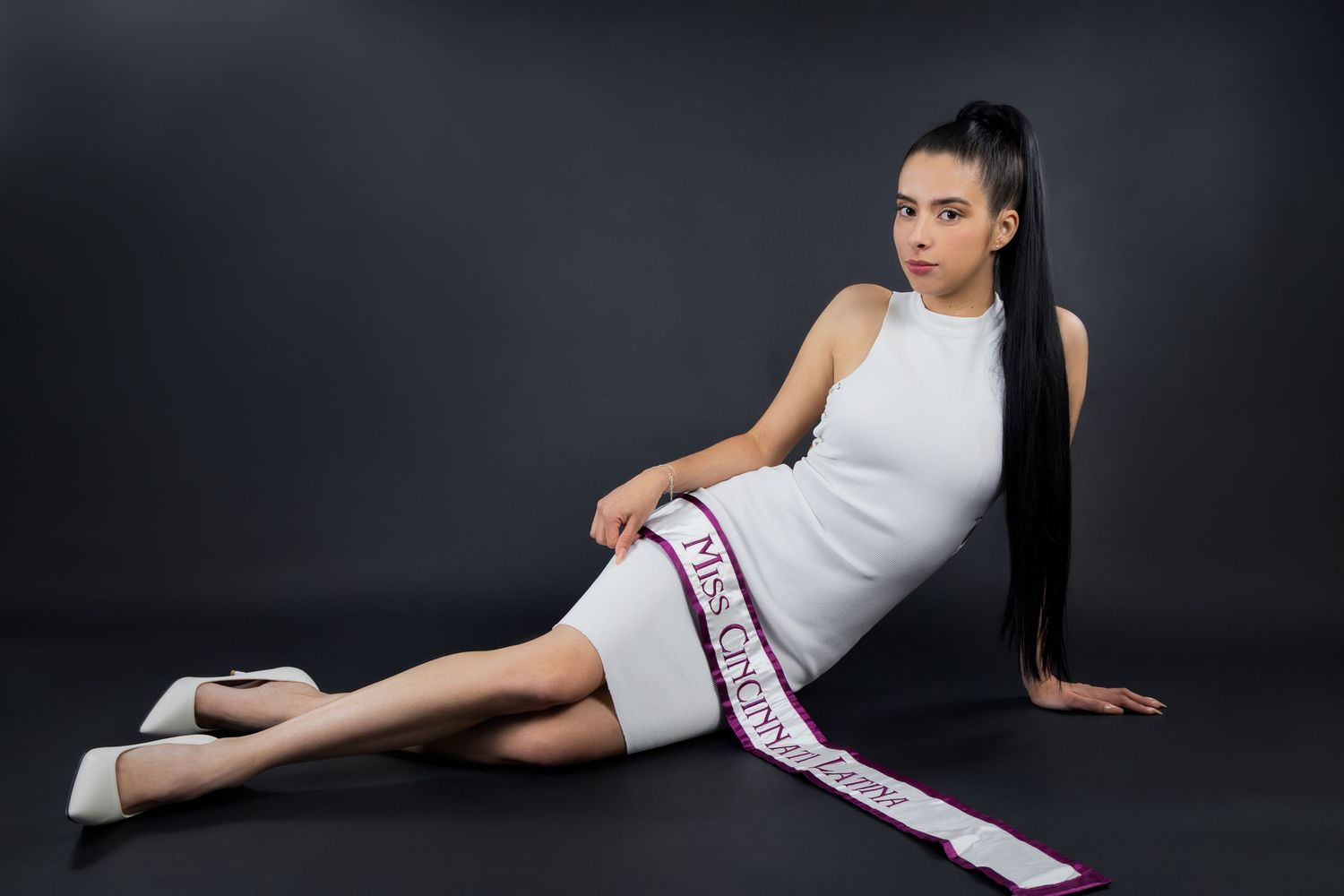 Young woman in white dress wearing a pageant sash poses gracefully on floor against dark gray background.