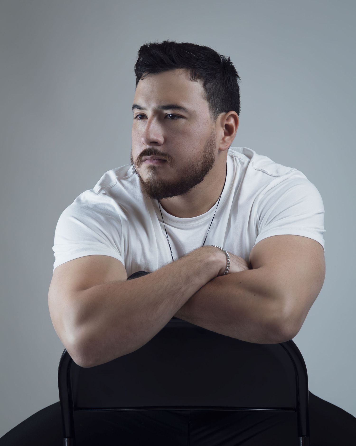 Muscular man in white t-shirt sits arms crossed on chair, looking away thoughtfully against gray background.