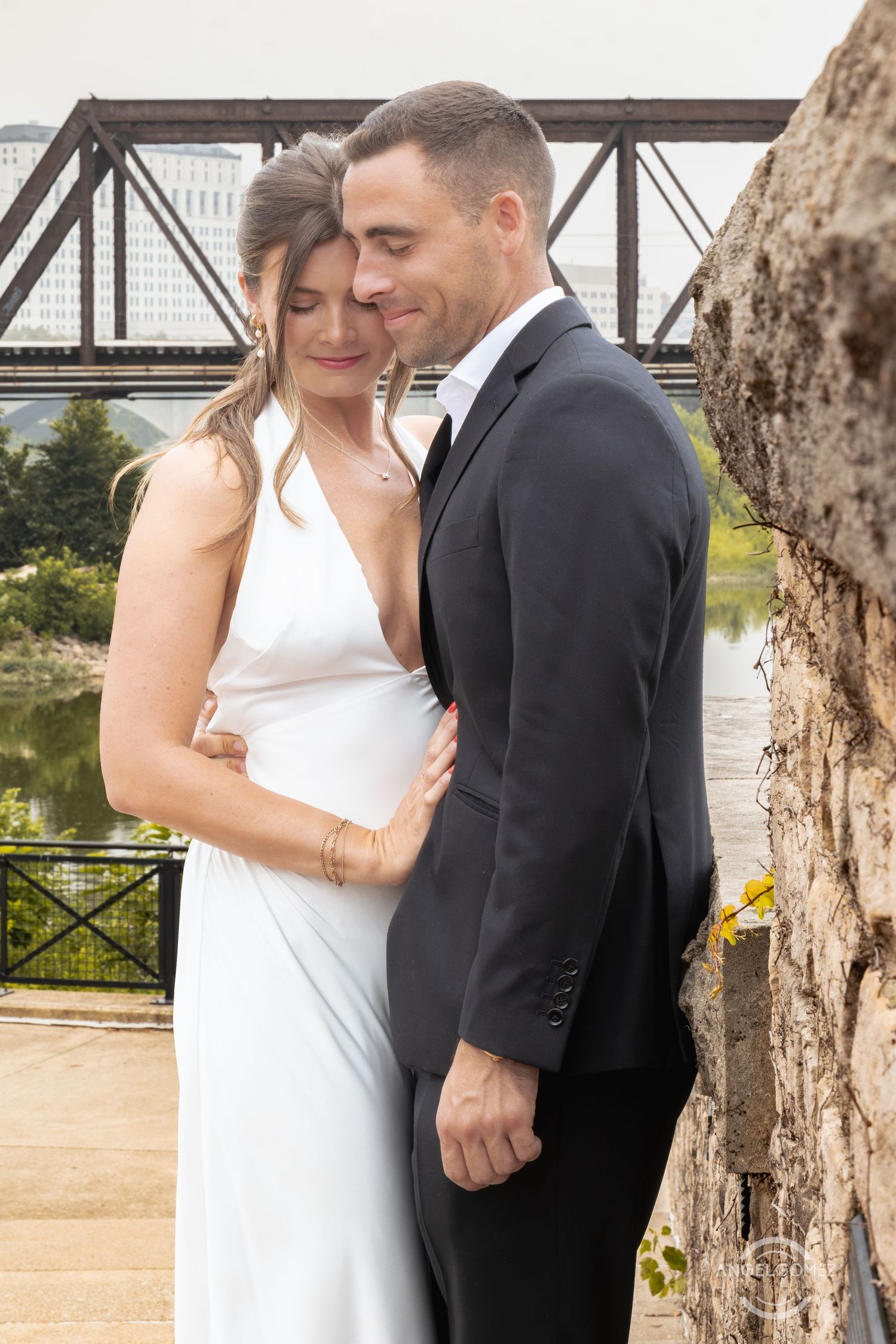 Bride in white gown and groom in black tuxedo embrace near stone wall with river bridge backdrop.