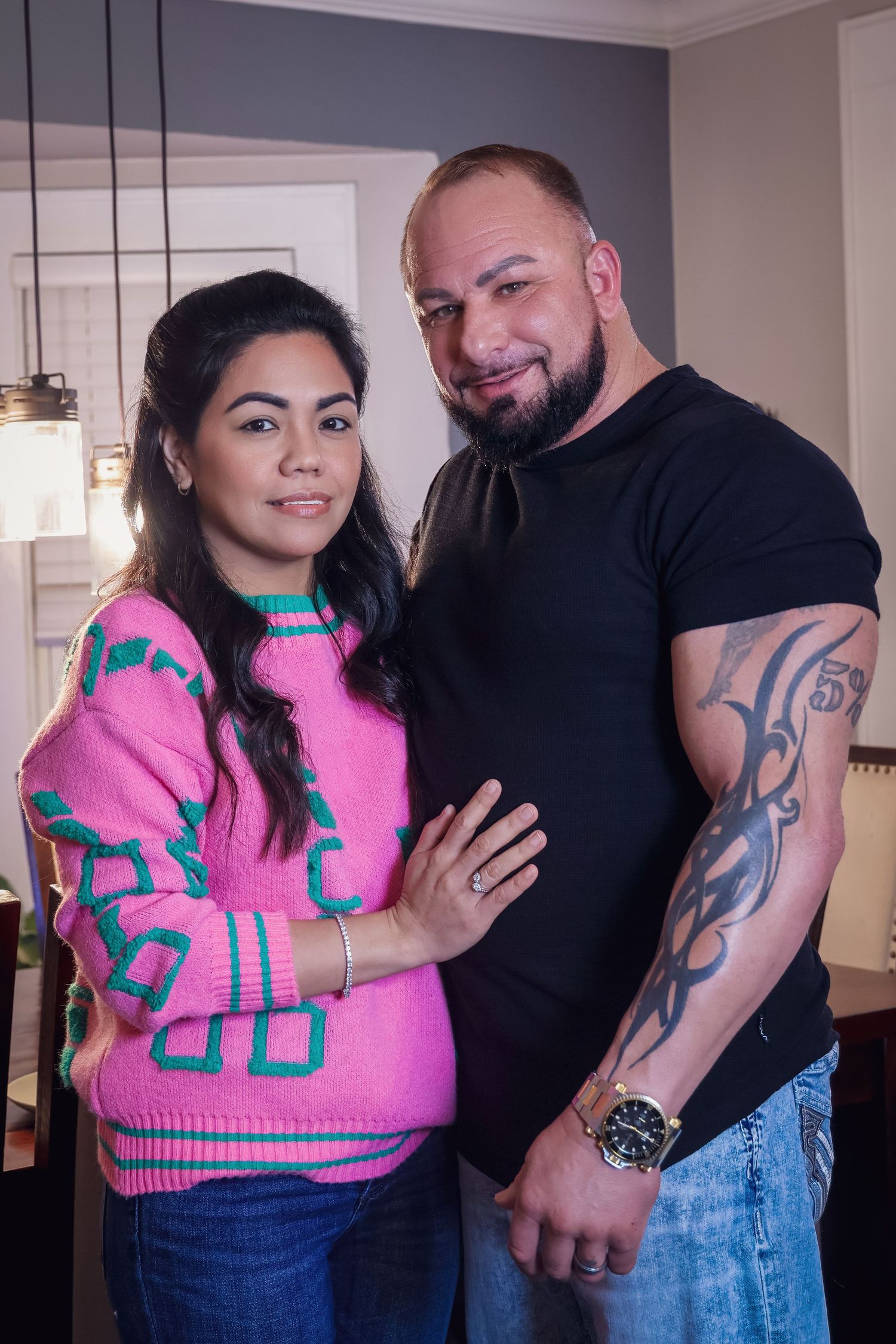 A couple posing together indoors against a grey wall with hanging pendant lights.