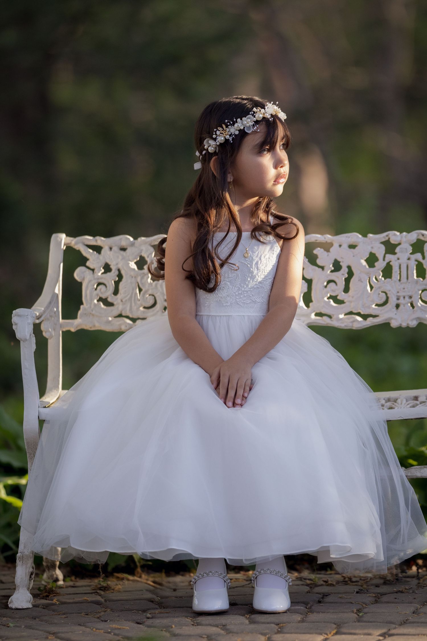 Young girl in white tulle dress and floral crown sits on ornate white bench outdoors.