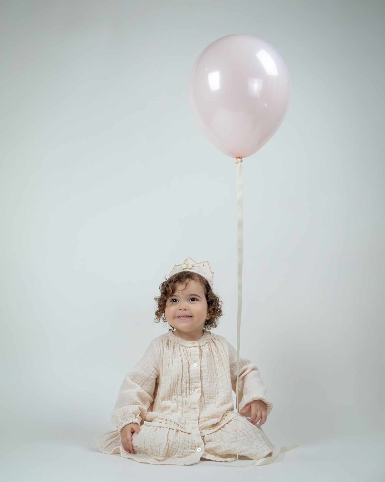 A smiling toddler in a cream dress and crown sits cross-legged holding a pink balloon on a white background.