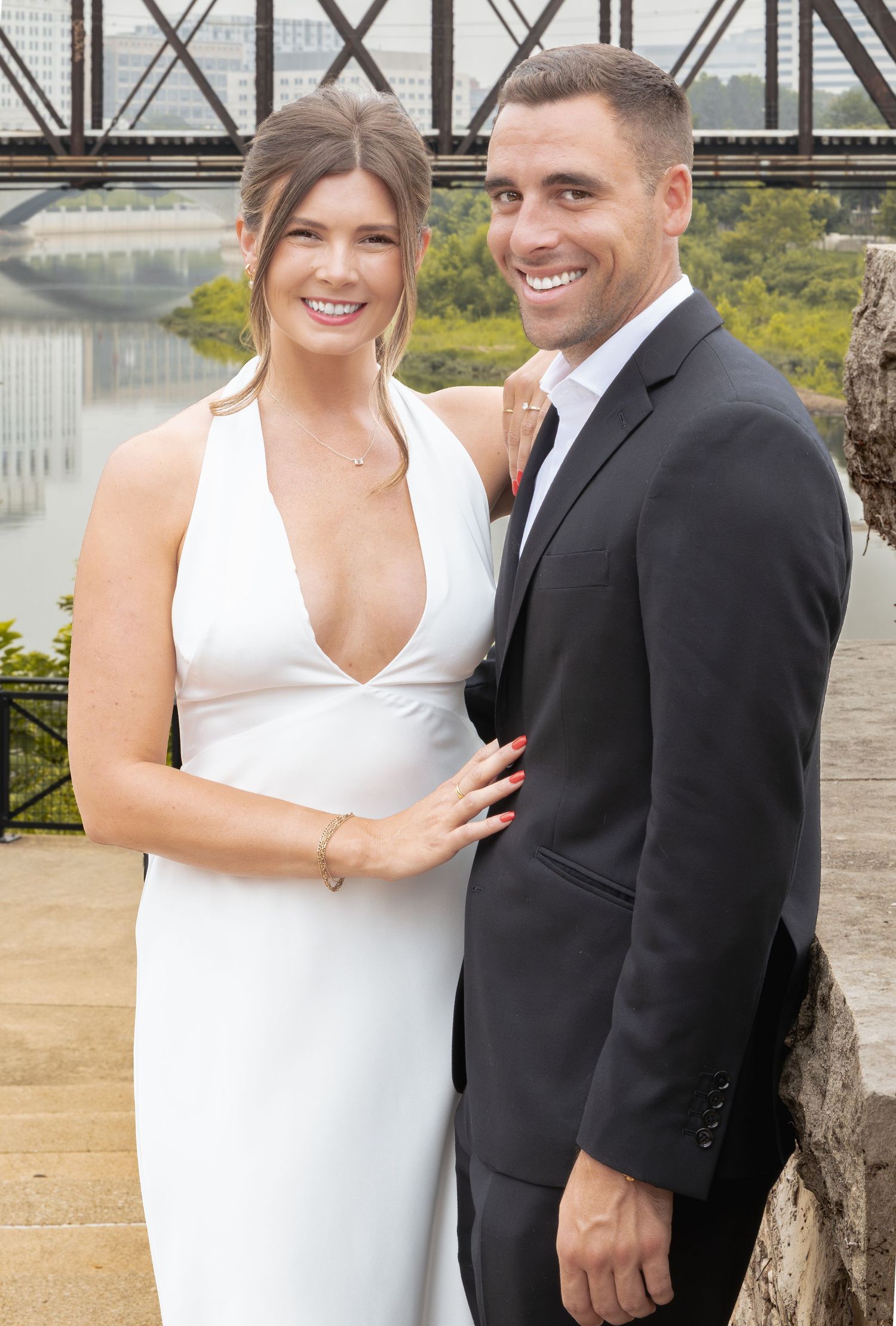 Couple posing outdoors near a river, woman in white gown and man in black suit, smiling together.