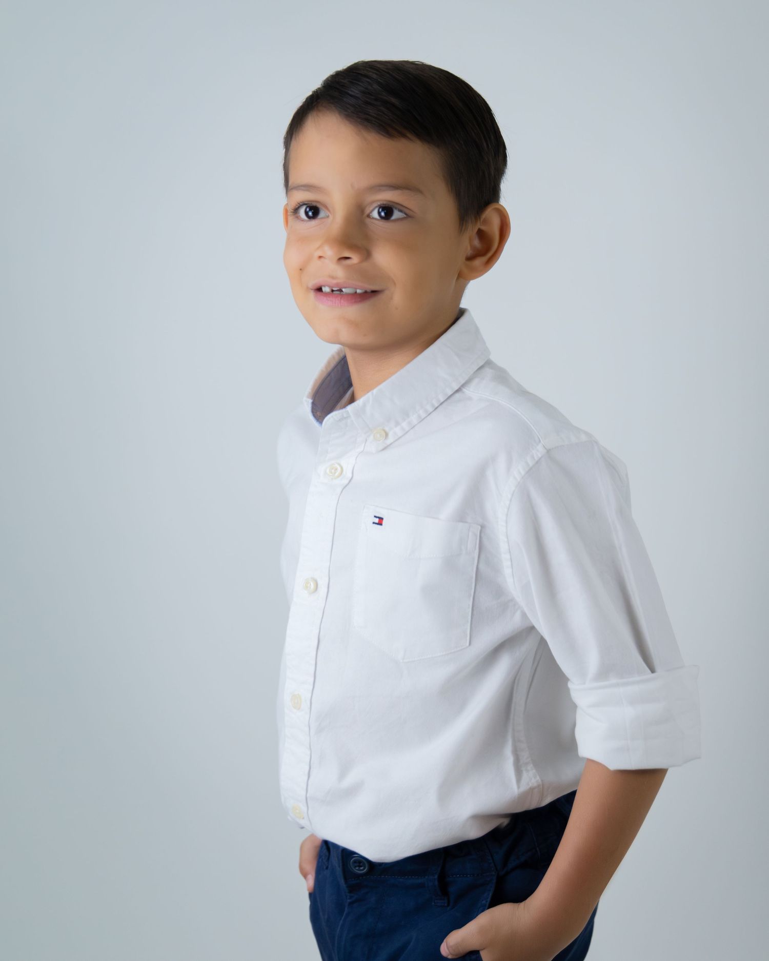 Young boy in white Tommy Hilfiger shirt and navy pants poses confidently against light gray background.