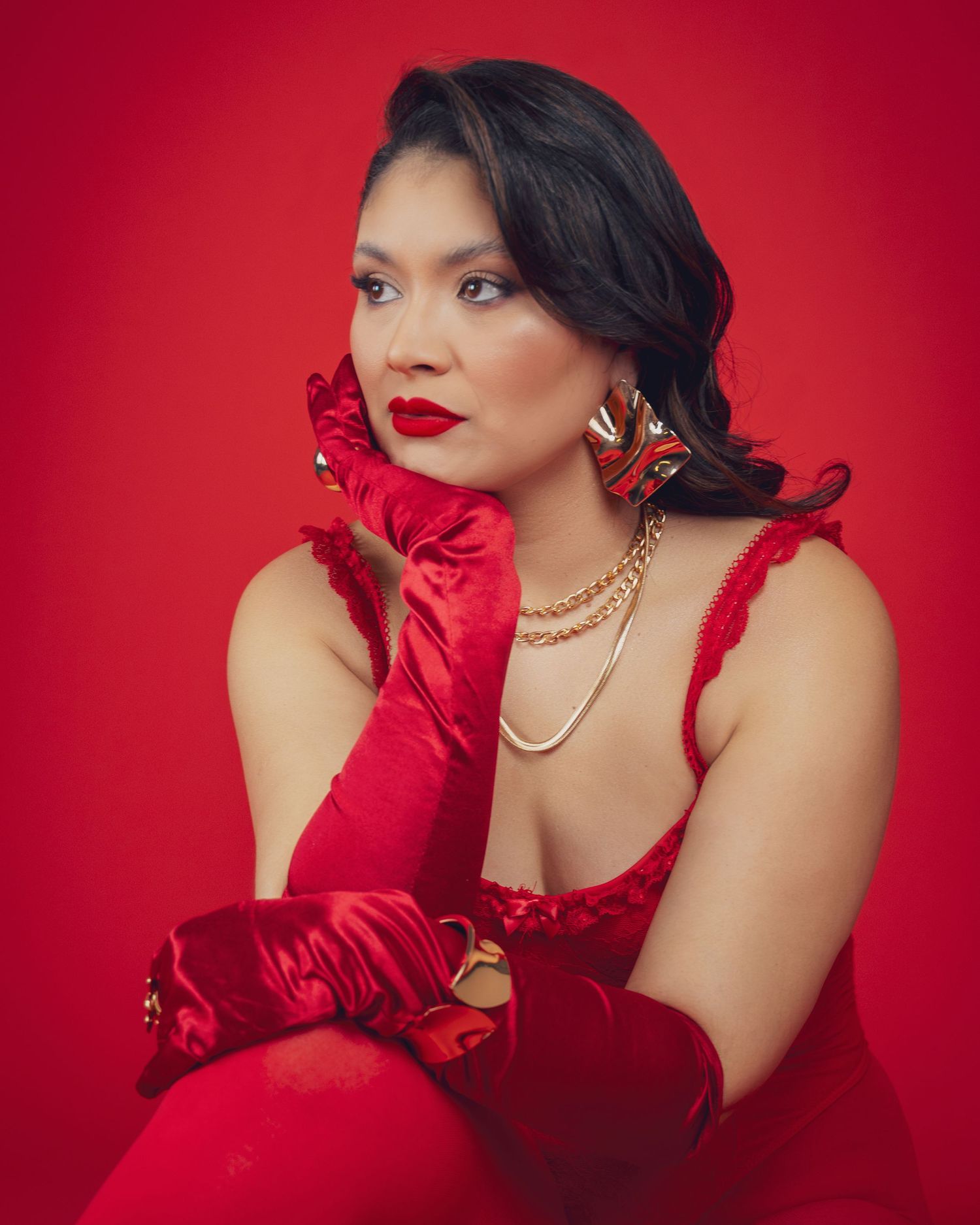 Woman in red satin dress and long gloves with gold jewelry poses elegantly against a red background.