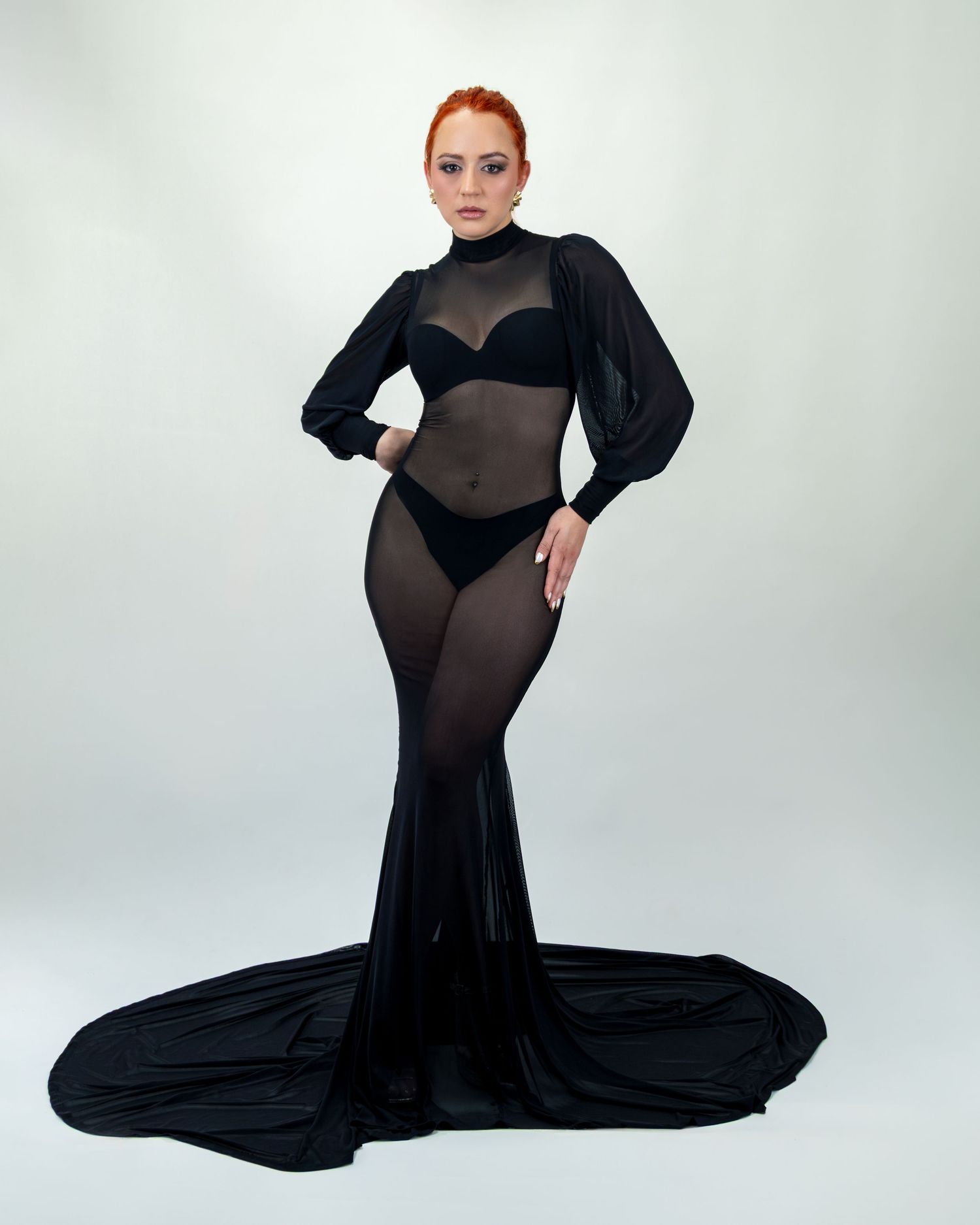 A woman with red hair poses in a sheer black mermaid gown with long sleeves and a dramatic train.
