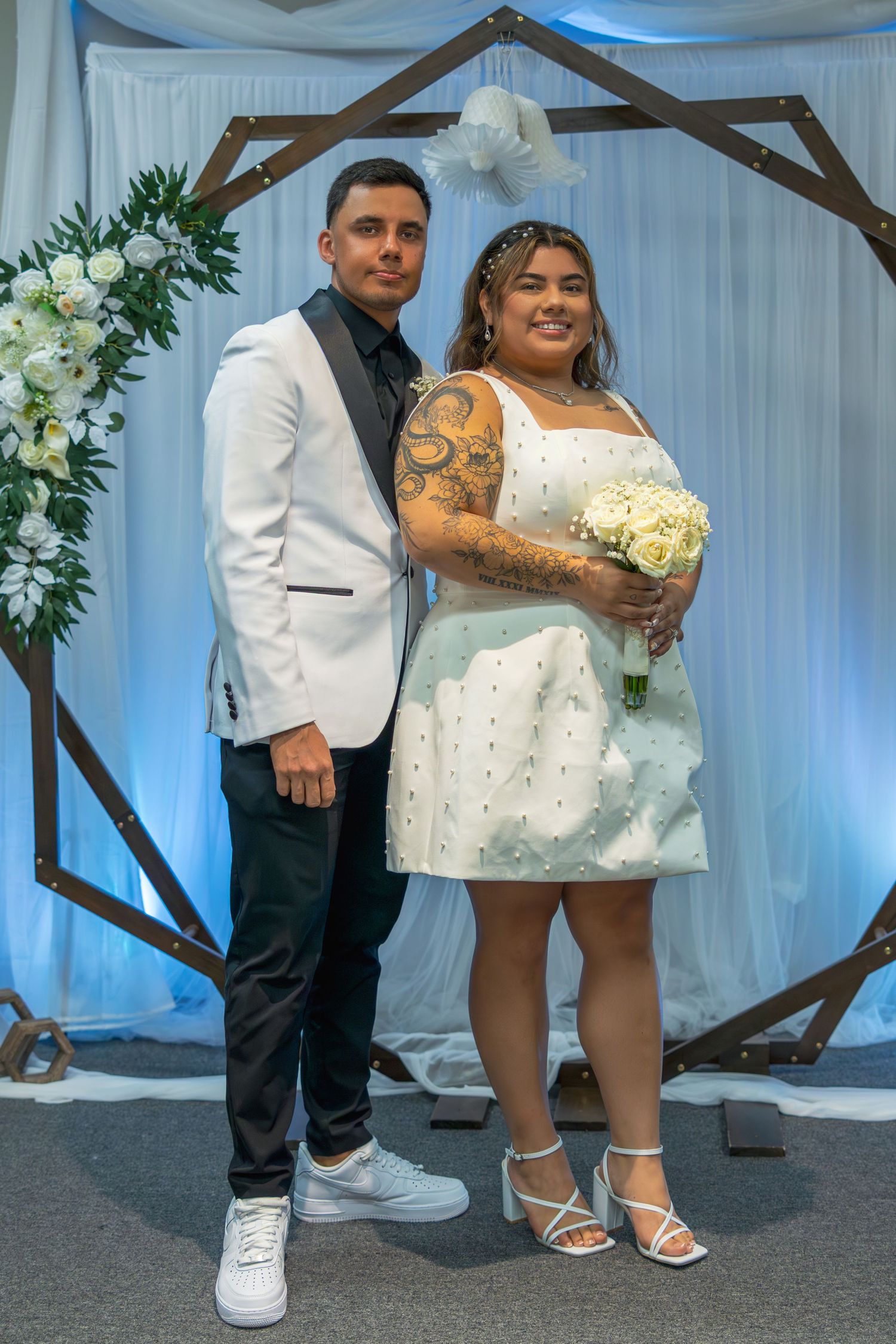 Couple posing at wedding altar, groom in white blazer, bride in short white dress holding bouquet, geometric arch backdrop.