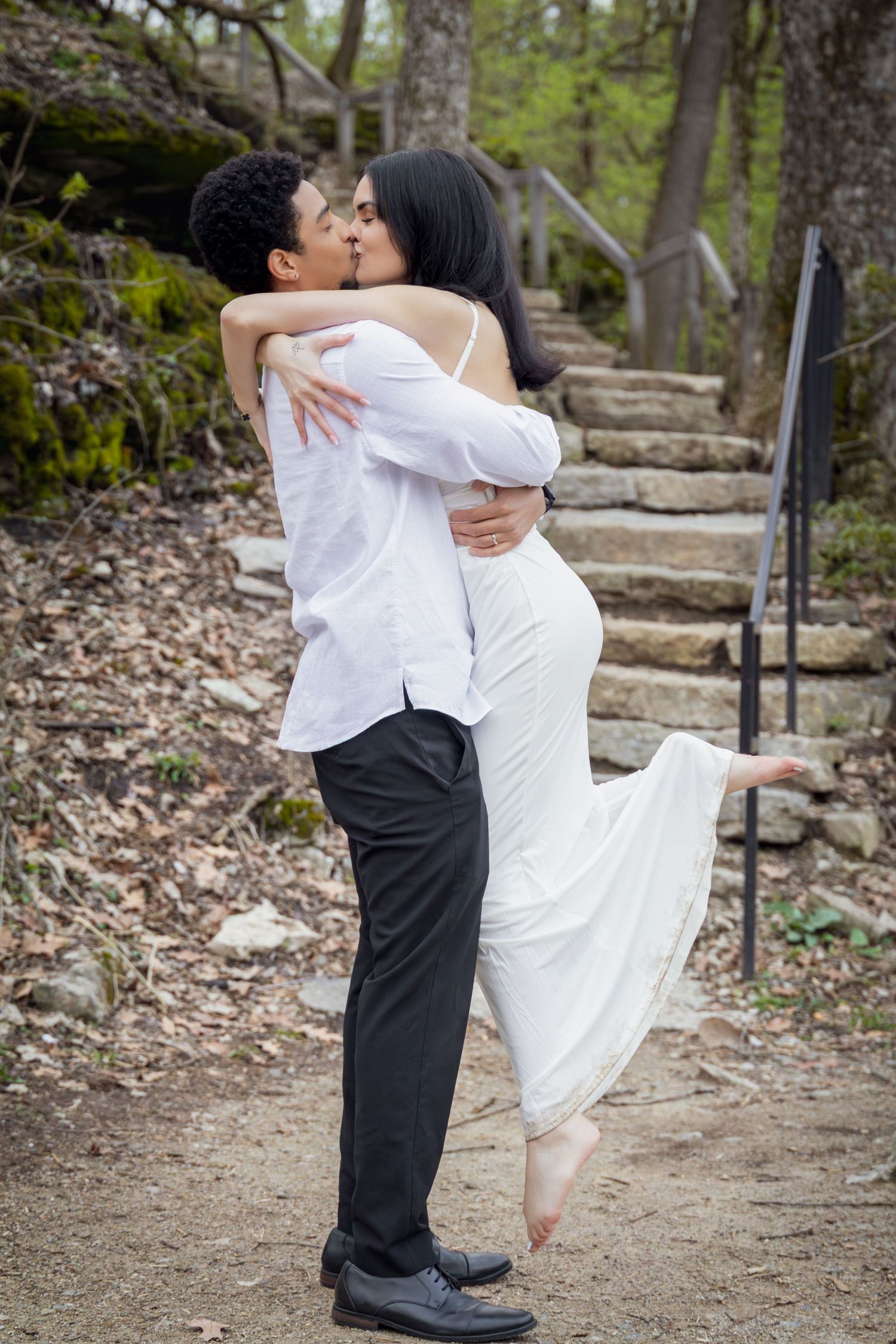 A couple shares a romantic kiss outdoors, man in white shirt and black pants lifting woman in white dress near stone steps.