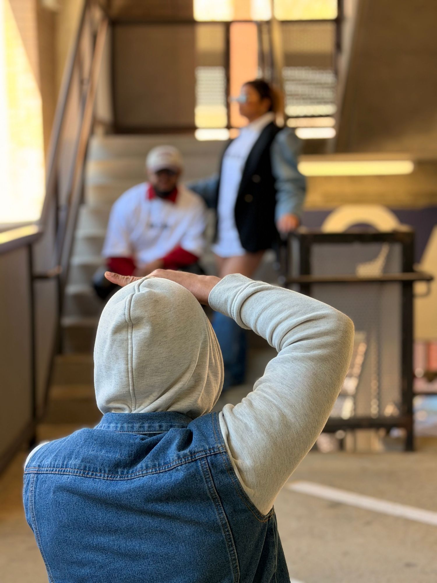 Person in denim jacket and hoodie photographing two people on stairs in an indoor setting.
