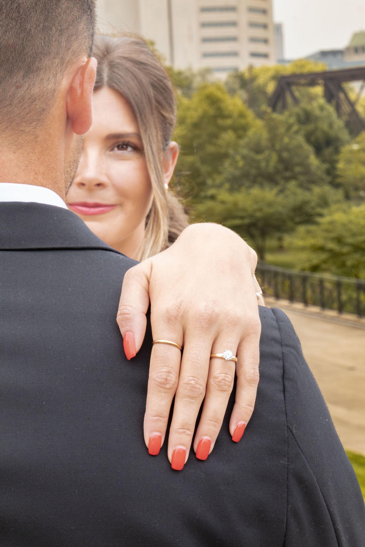 Woman showing off engagement ring with red nails over man's shoulder in formal wear outdoors.