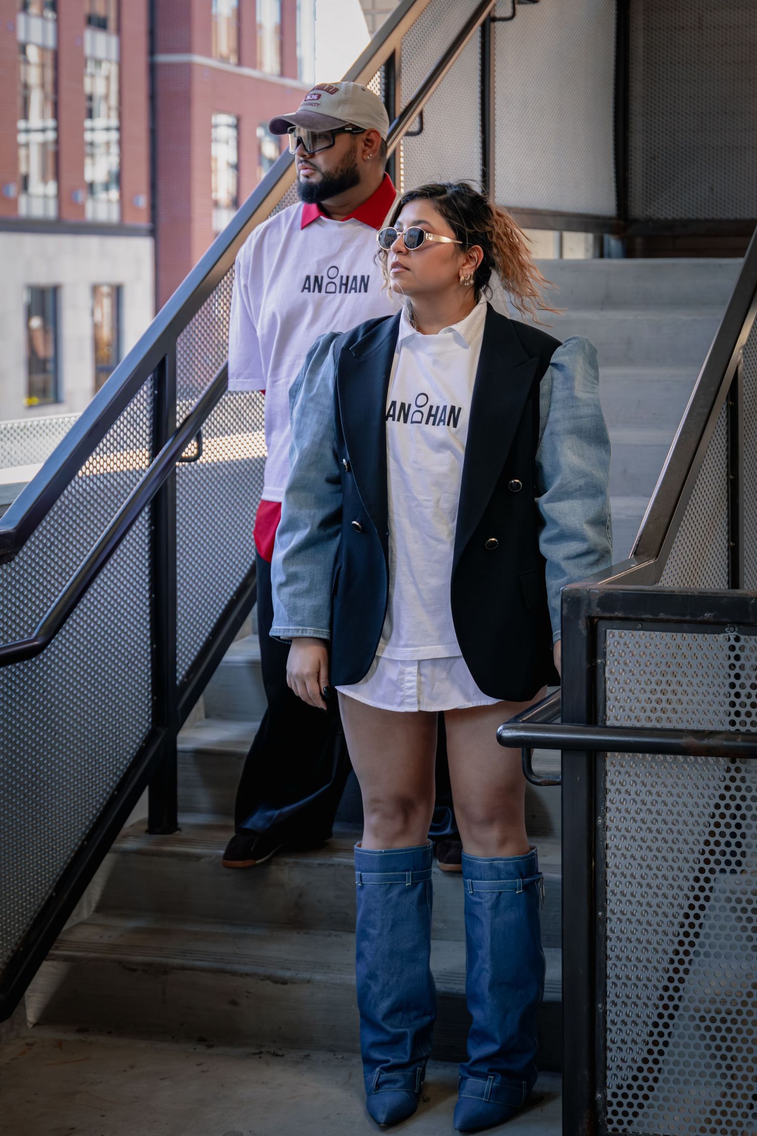 Two stylish people wearing matching AN2HAN branded white tees and navy blazers pose on urban stairs.