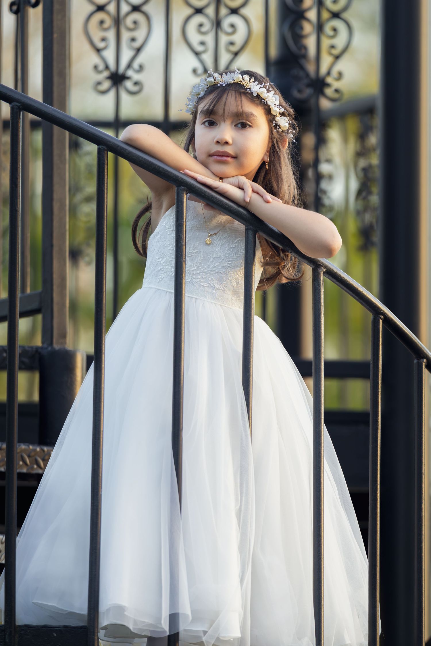 Young girl in white flower girl dress and floral crown posing gracefully on decorative iron staircase railing.