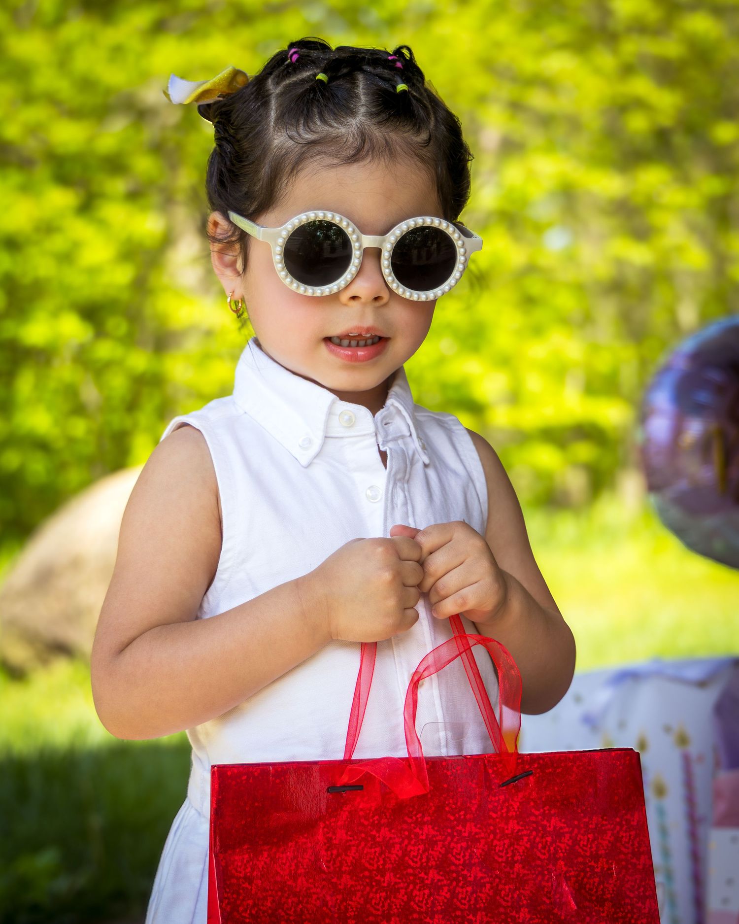 A cute little girl wearing round white sunglasses holds a red gift bag outdoors in a lush green setting.