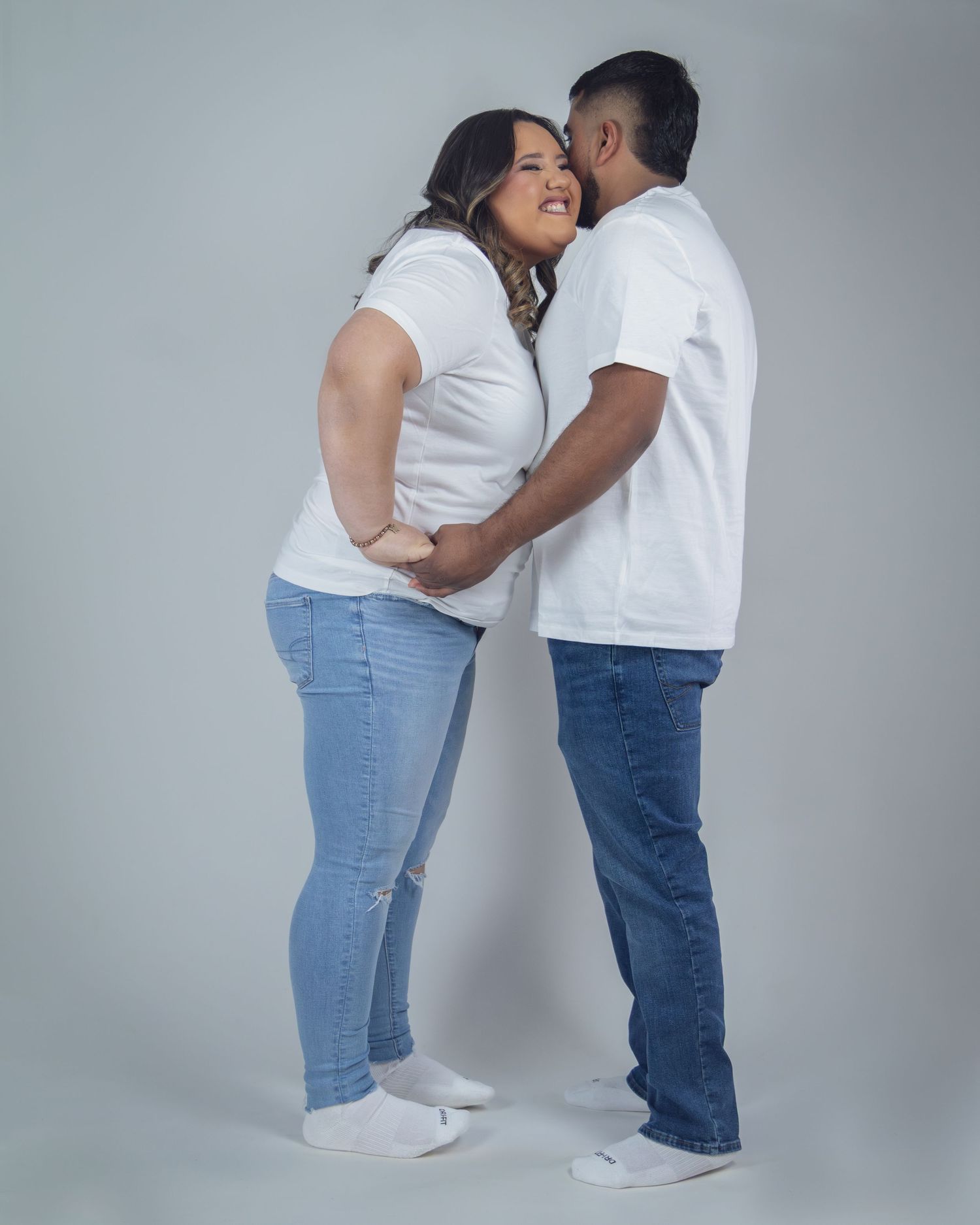 A loving couple in matching white t-shirts and jeans share a tender moment, smiling and embracing against a gray background.