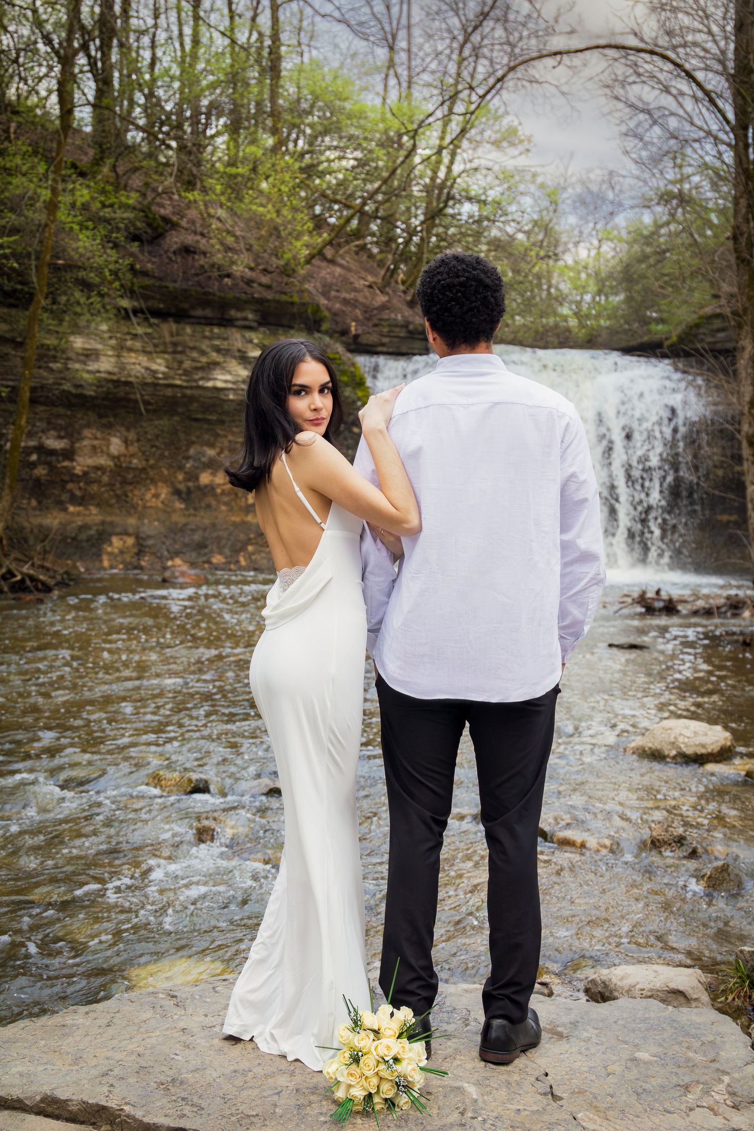 Bride in backless white gown and groom stand on rocks before a waterfall, holding yellow flowers.