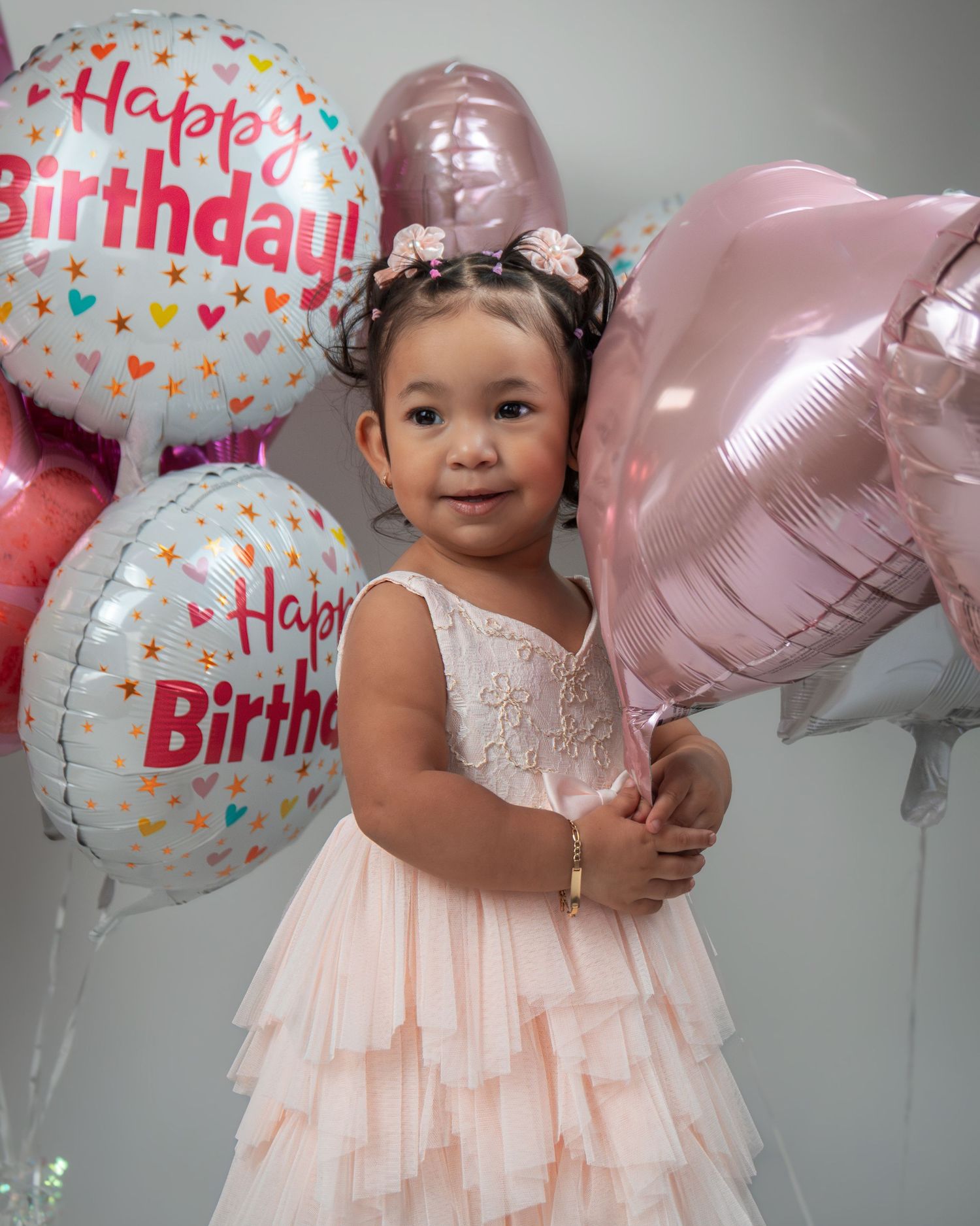 A young toddler in a ruffled peach-colored dress stands among pink and white birthday balloons during a celebration.