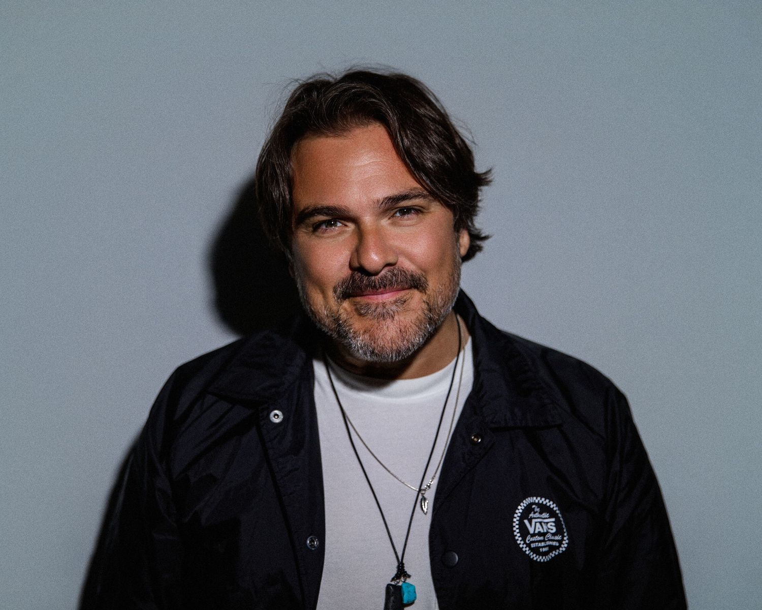 Man with brown hair and beard wearing a Vans jacket and layered necklaces poses against a gray background.