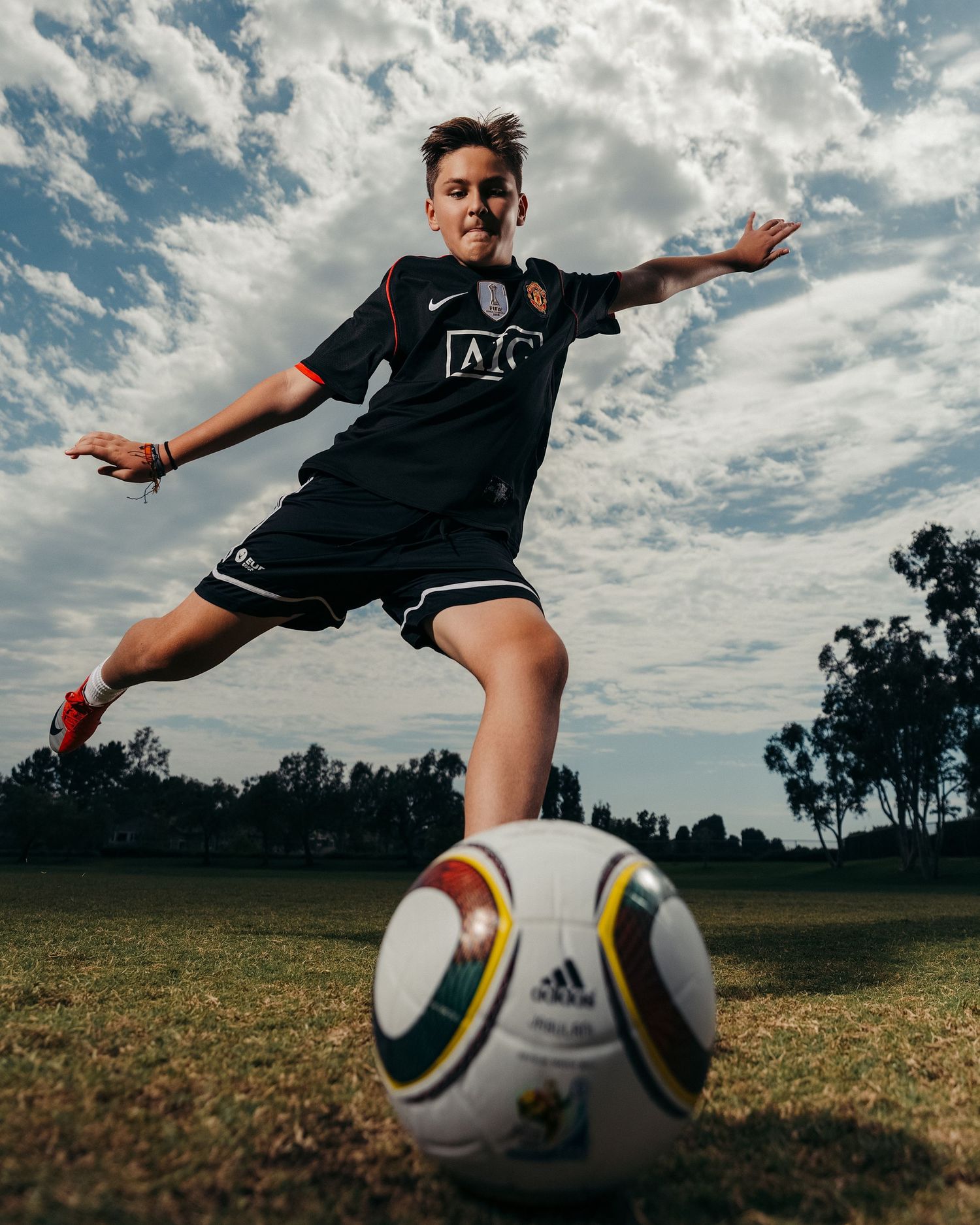 Young soccer player in black Manchester United jersey kicking an Adidas ball on a grass field under a cloudy sky.