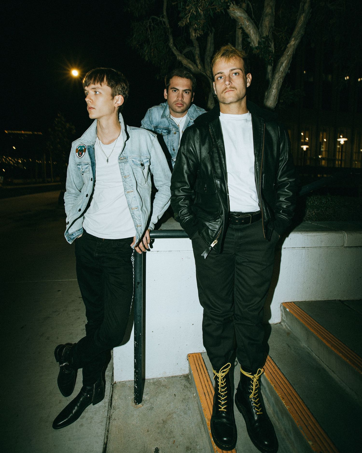 Three young men in stylish jackets and black pants pose outdoors at night near a bench and trees.