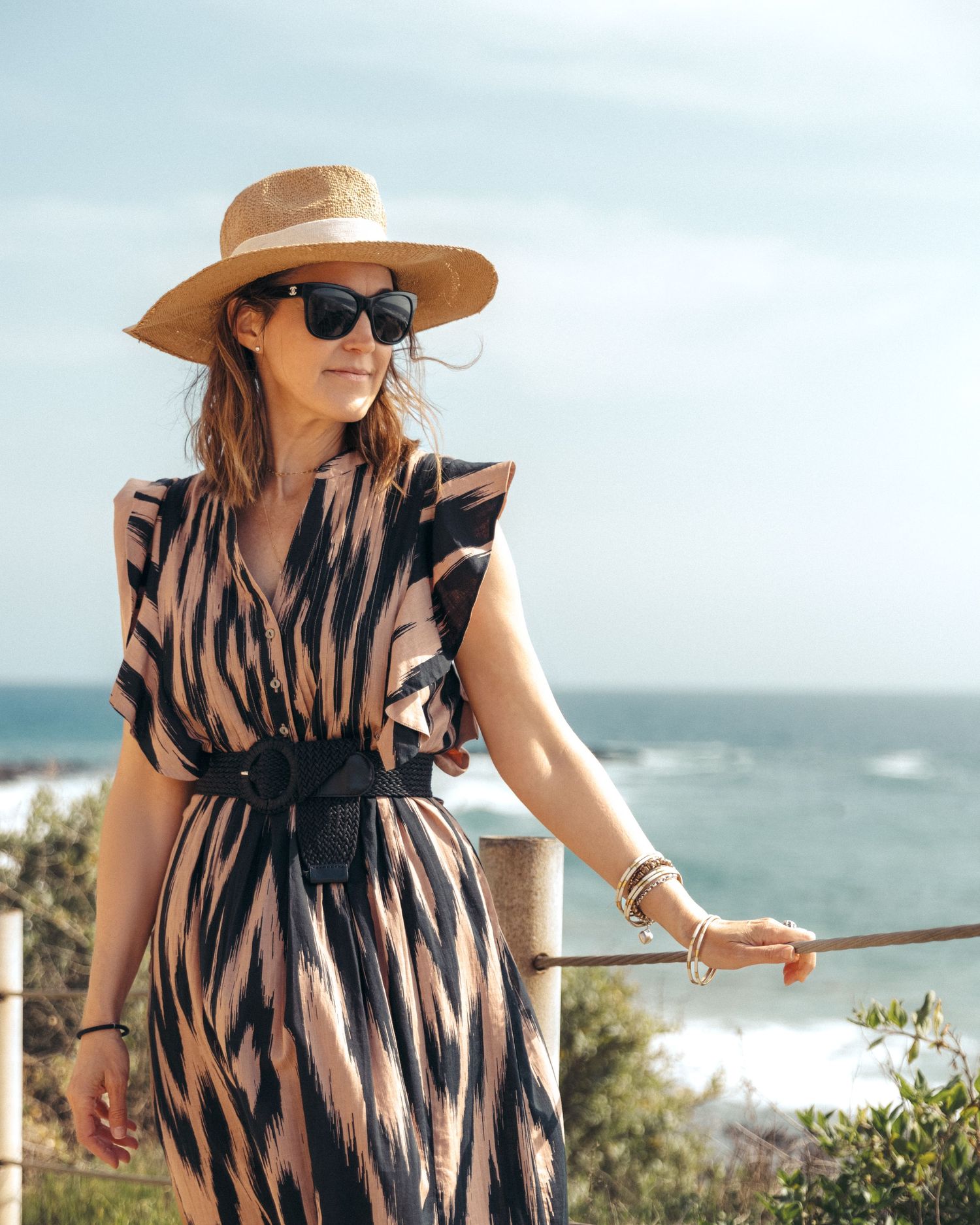 A woman in a striped summer dress and sun hat stands by a coastal railing overlooking the ocean on a sunny day.