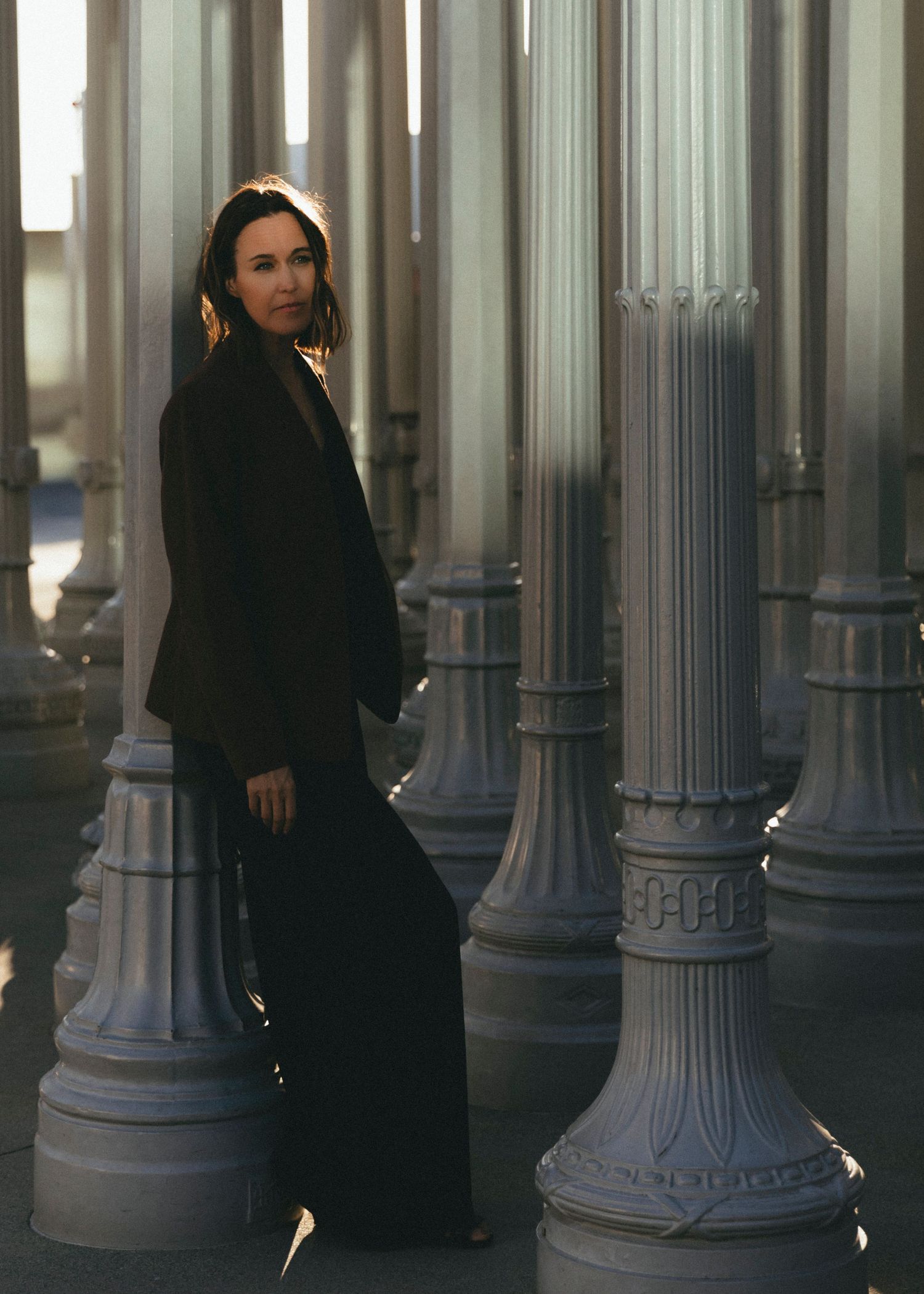 A person in black clothing stands among ornate white columns in an architectural space with dramatic lighting.