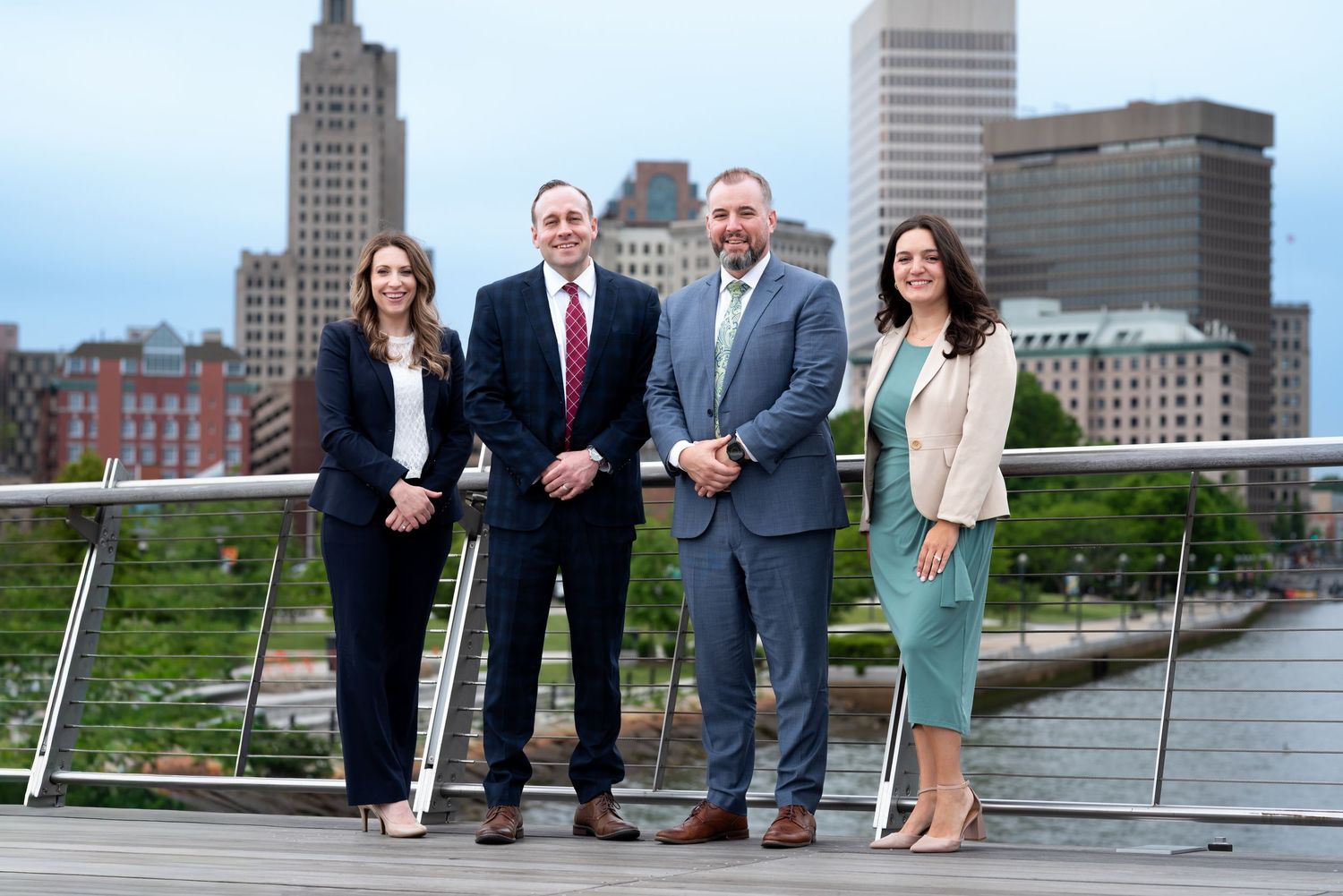 Four business professionals smiling on a waterfront terrace with a city skyline backdrop.