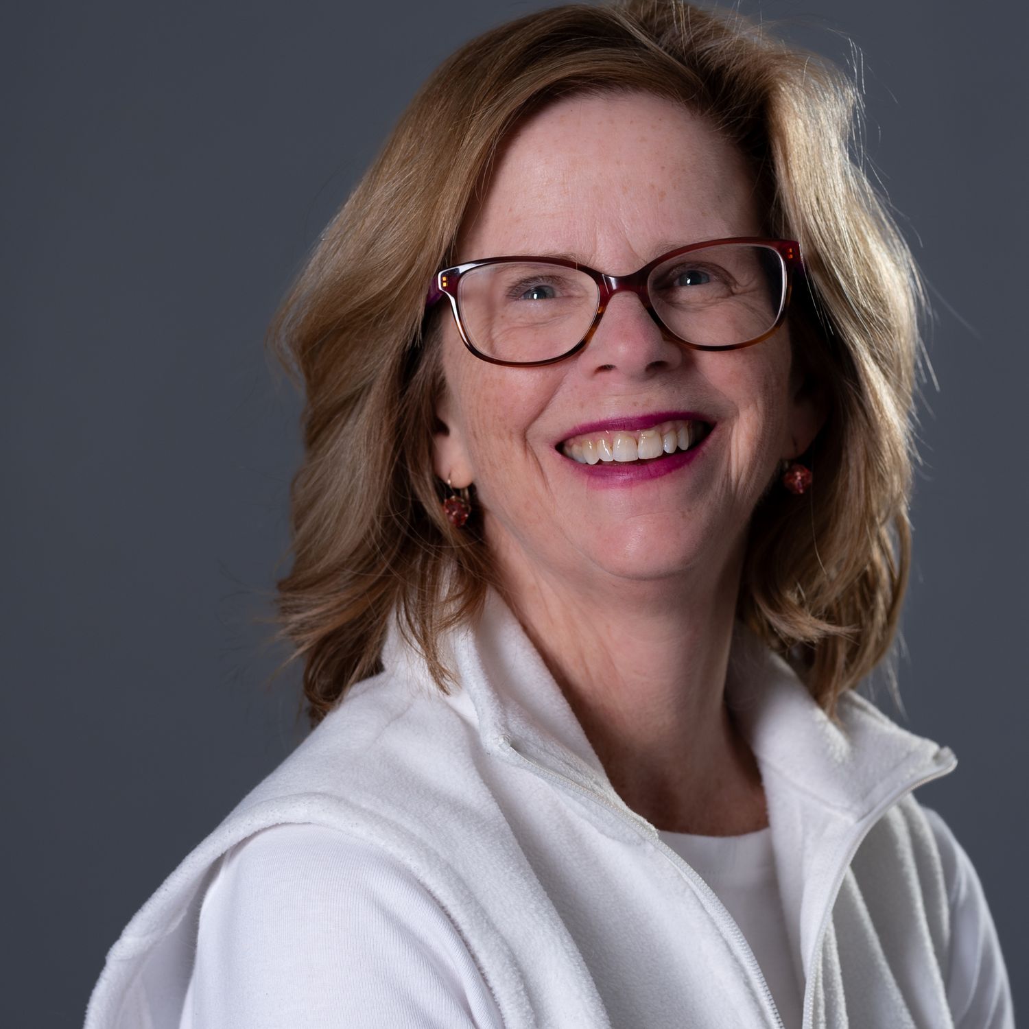Smiling middle-aged woman with red glasses and white jacket against gray background.